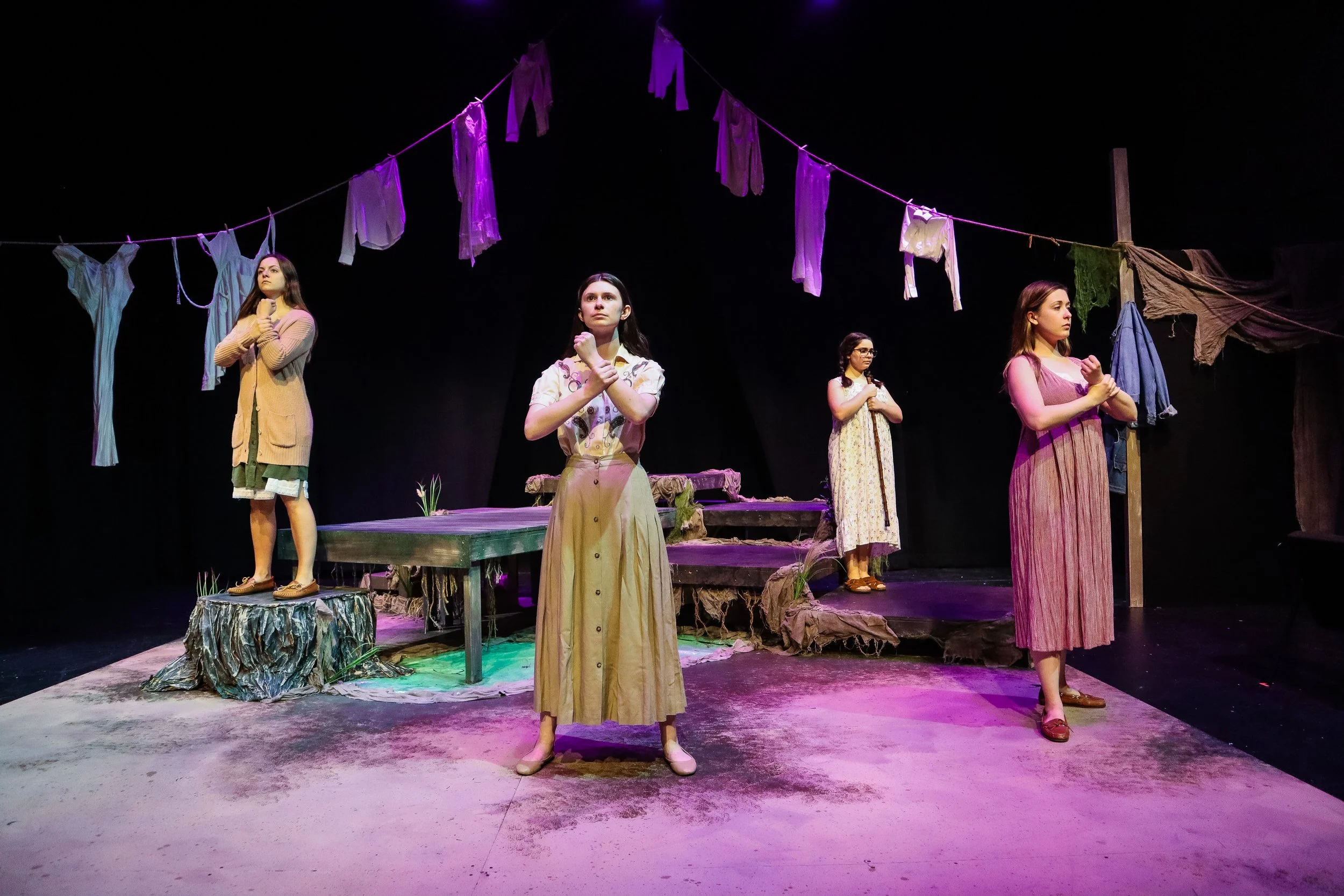Four women are on stage, two standing and two sitting, with laundry hanging above them on a line, set in a theatrical scene with a rustic, outdoor backdrop.