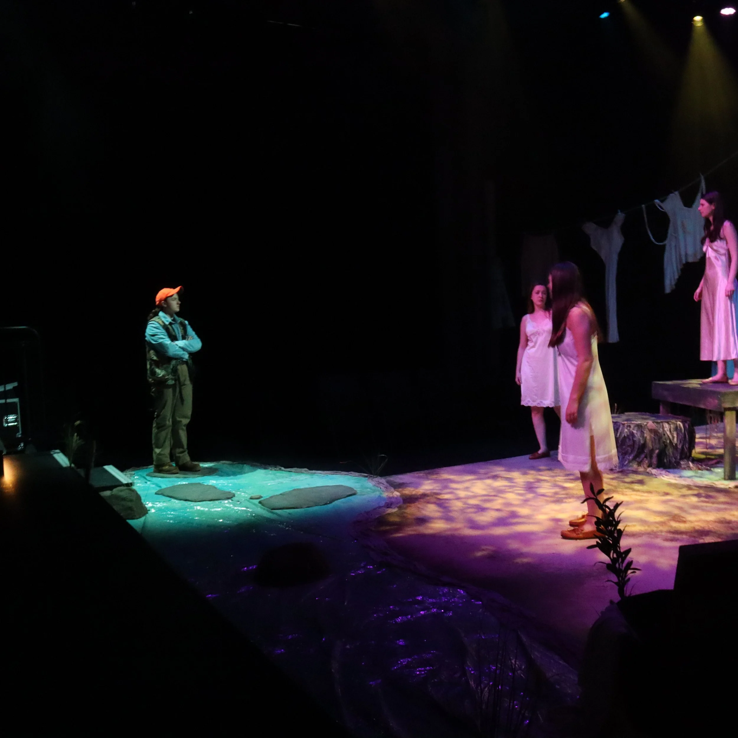 Stage scene from a theatrical performance with several actors dressed in costumes. A woman in a pink dress stands on the right, a girl in a pink shirt with orange cap and blue jacket on the left, with other actors partially visible. Stage lighting cr