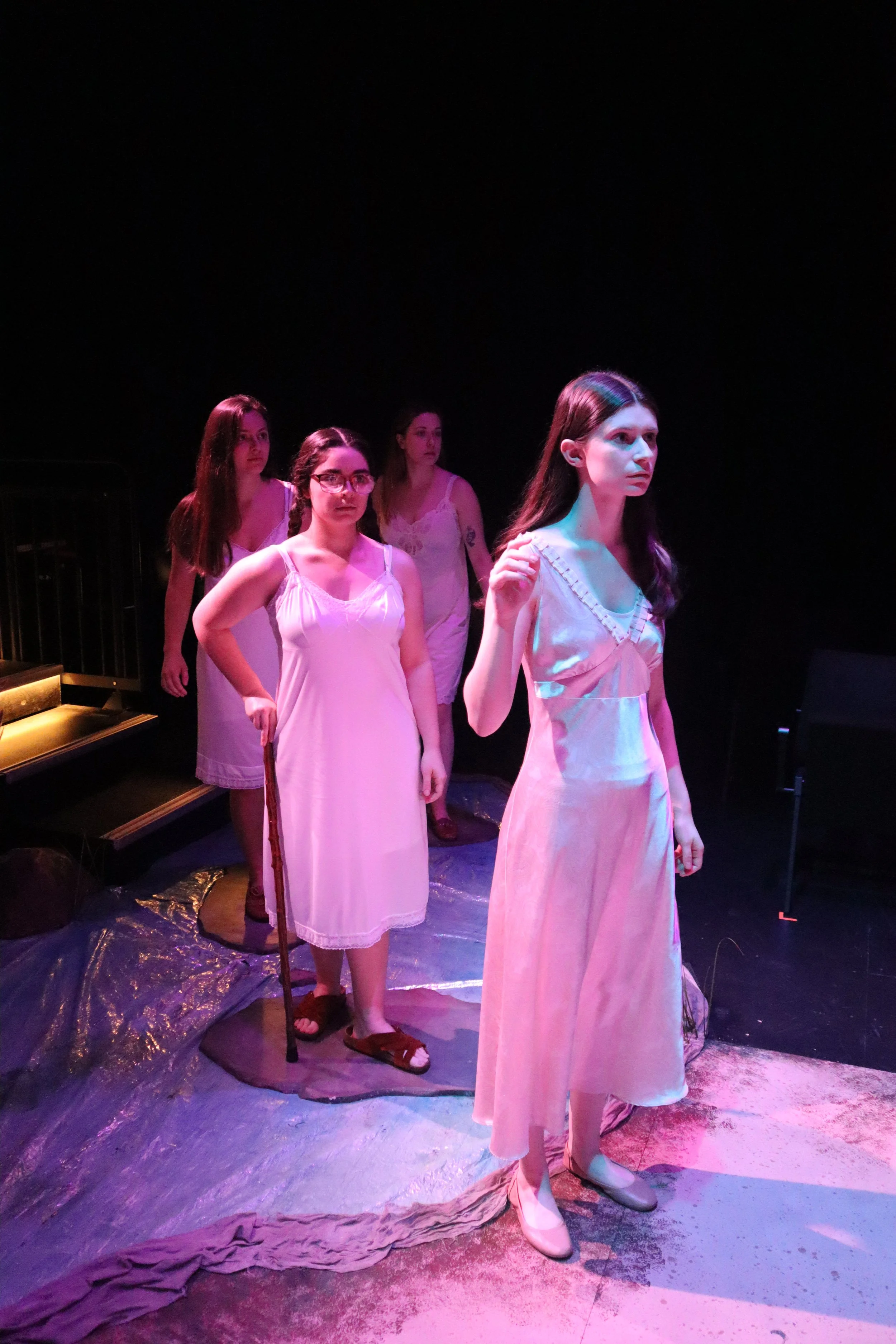 Four women standing on a stage, dressed in nightgown-style dresses, with dim, colored lighting. One woman in front holds a cane, appearing contemplative, while the others look on, some with serious expressions.