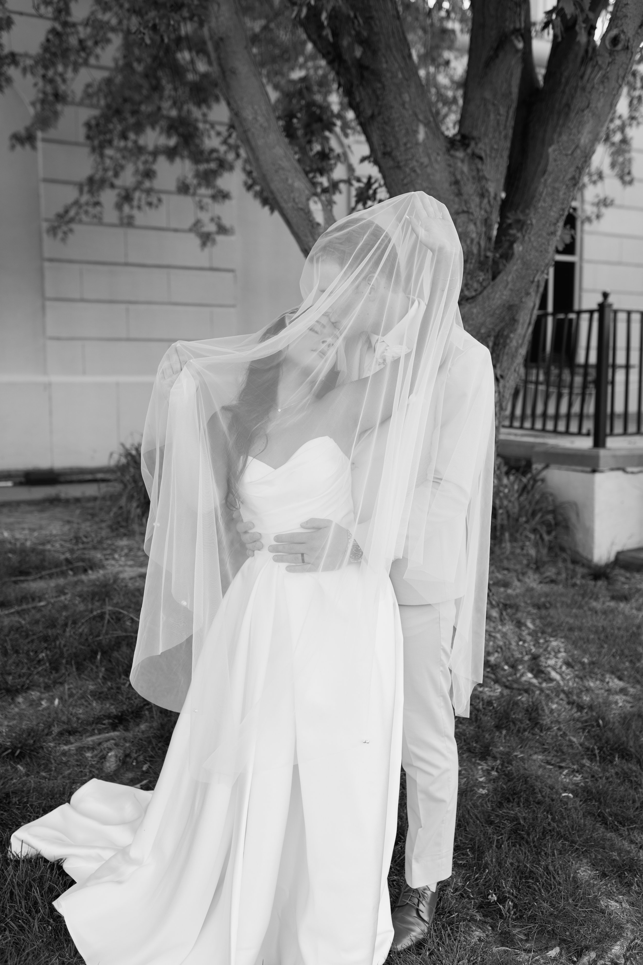A person dressed in a wedding gown with a veil covering their face, standing outdoors near a large tree and a building with a fence in the background.