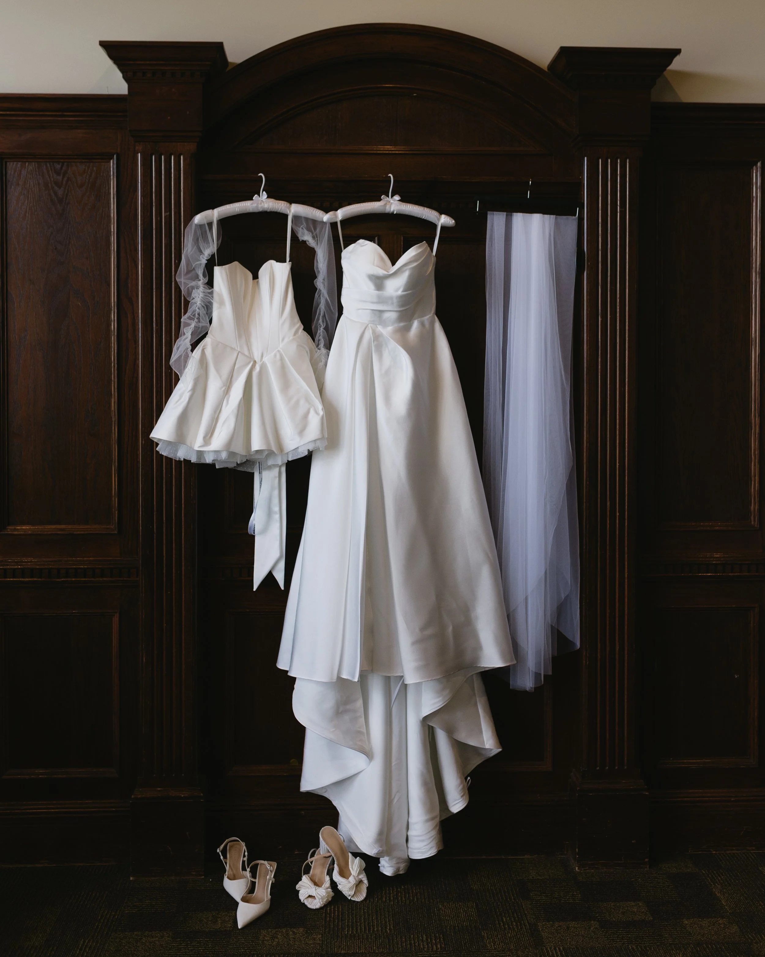 Two white wedding dresses hanging on a dark wooden wardrobe and two pairs of white shoes placed on the floor.