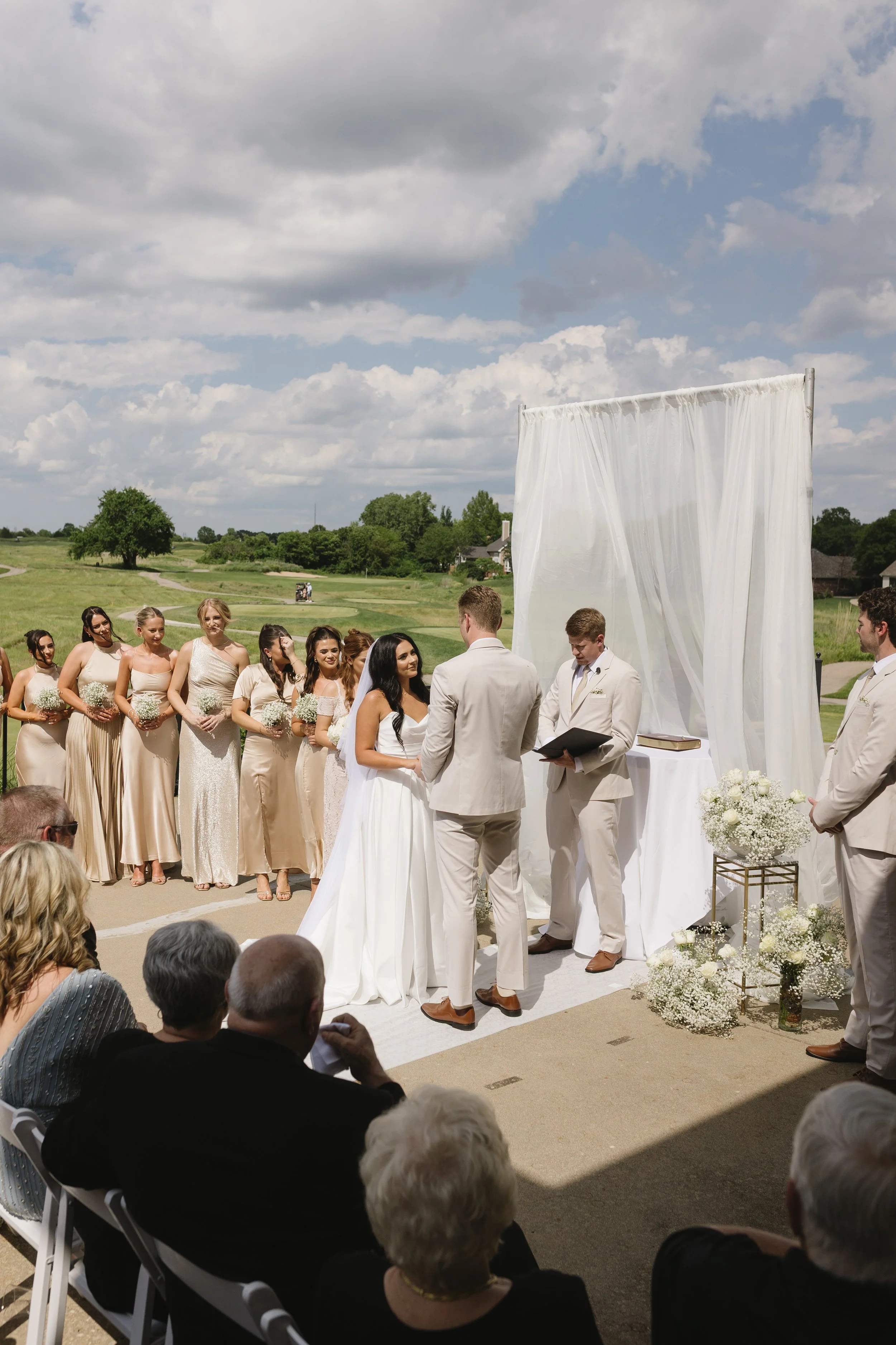 A wedding ceremony taking place outdoors on a bright day with blue skies and scattered clouds. The bride and groom stand facing each other under a white canopy with draped fabric, surrounded by bridesmaids dressed in beige and guests seated in chairs
