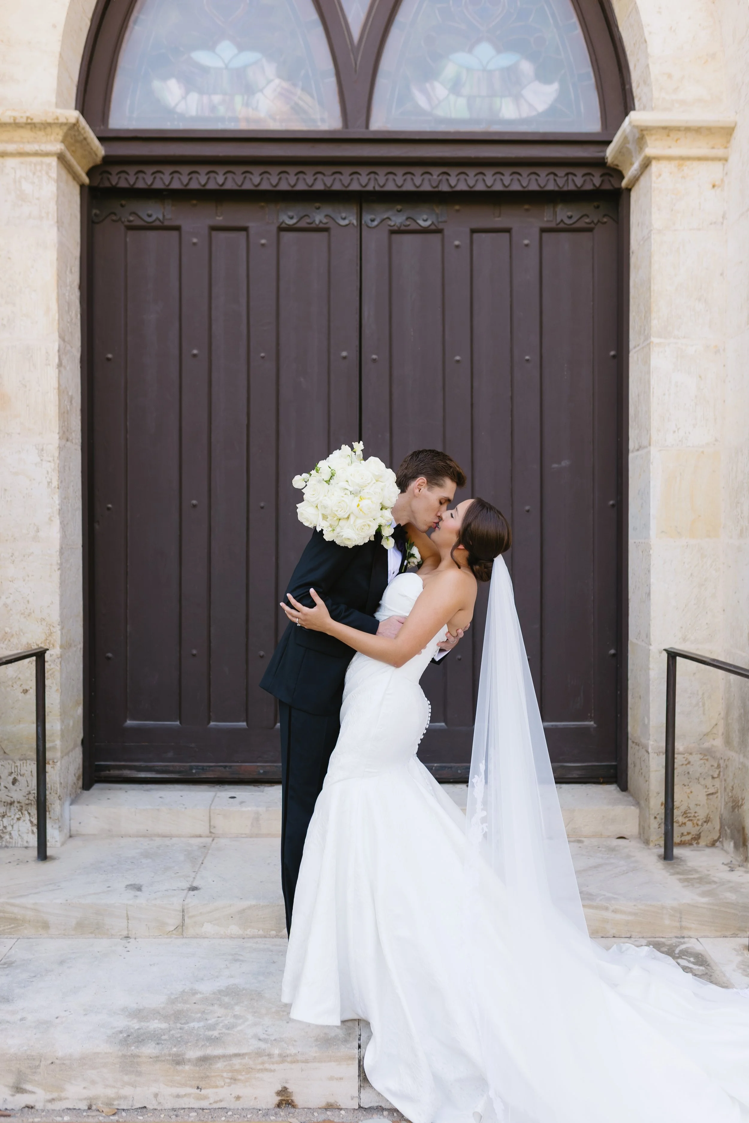 A bride and groom sharing a kiss outside a building with a large wooden door and stained glass window above. The groom is holding a bouquet of white roses, and the bride is wearing a strapless wedding gown with a long veil.