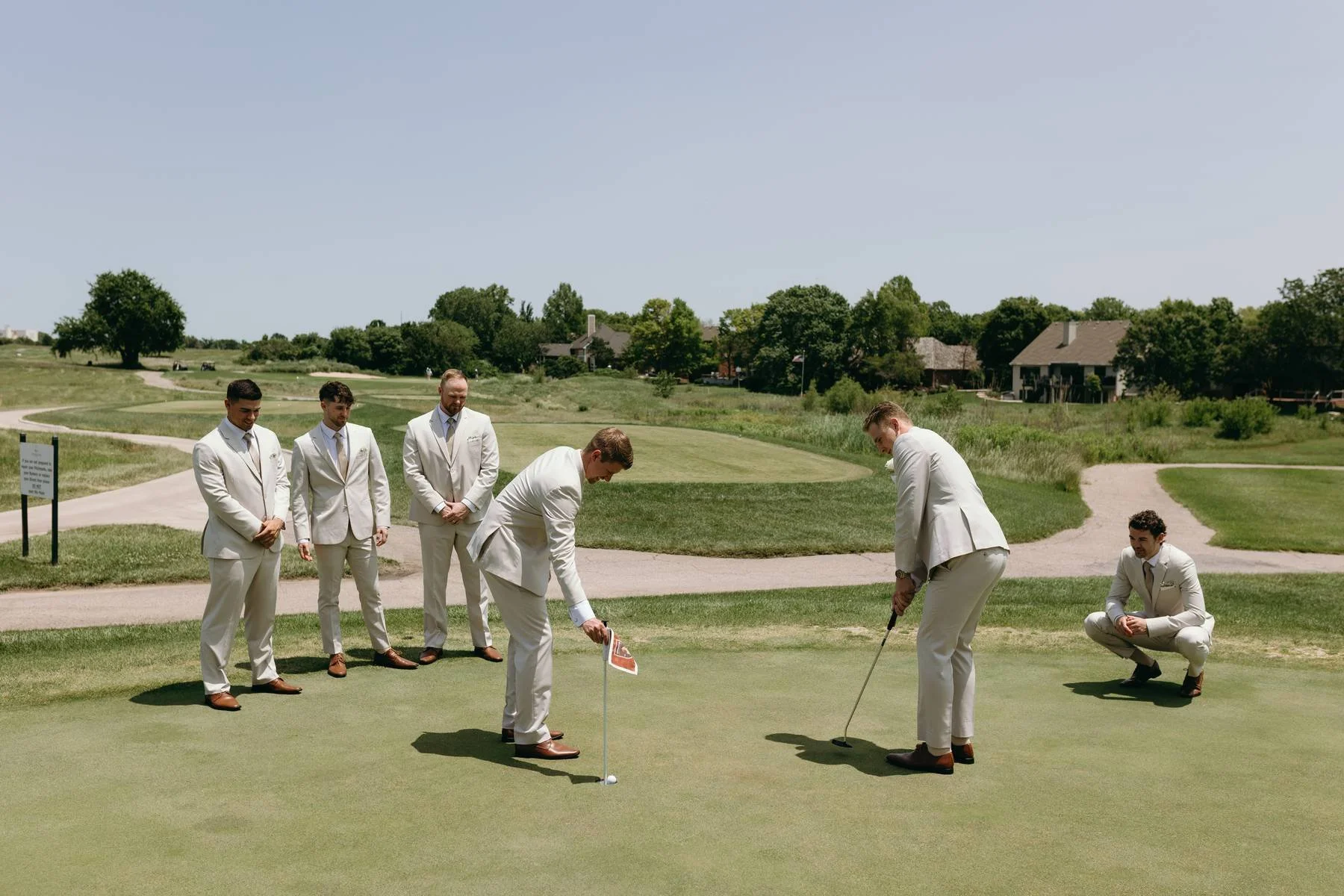 A group of six men dressed in white suits on a golf course, with three standing and three squatting, as one prepares to take a putt on the green under a clear blue sky.