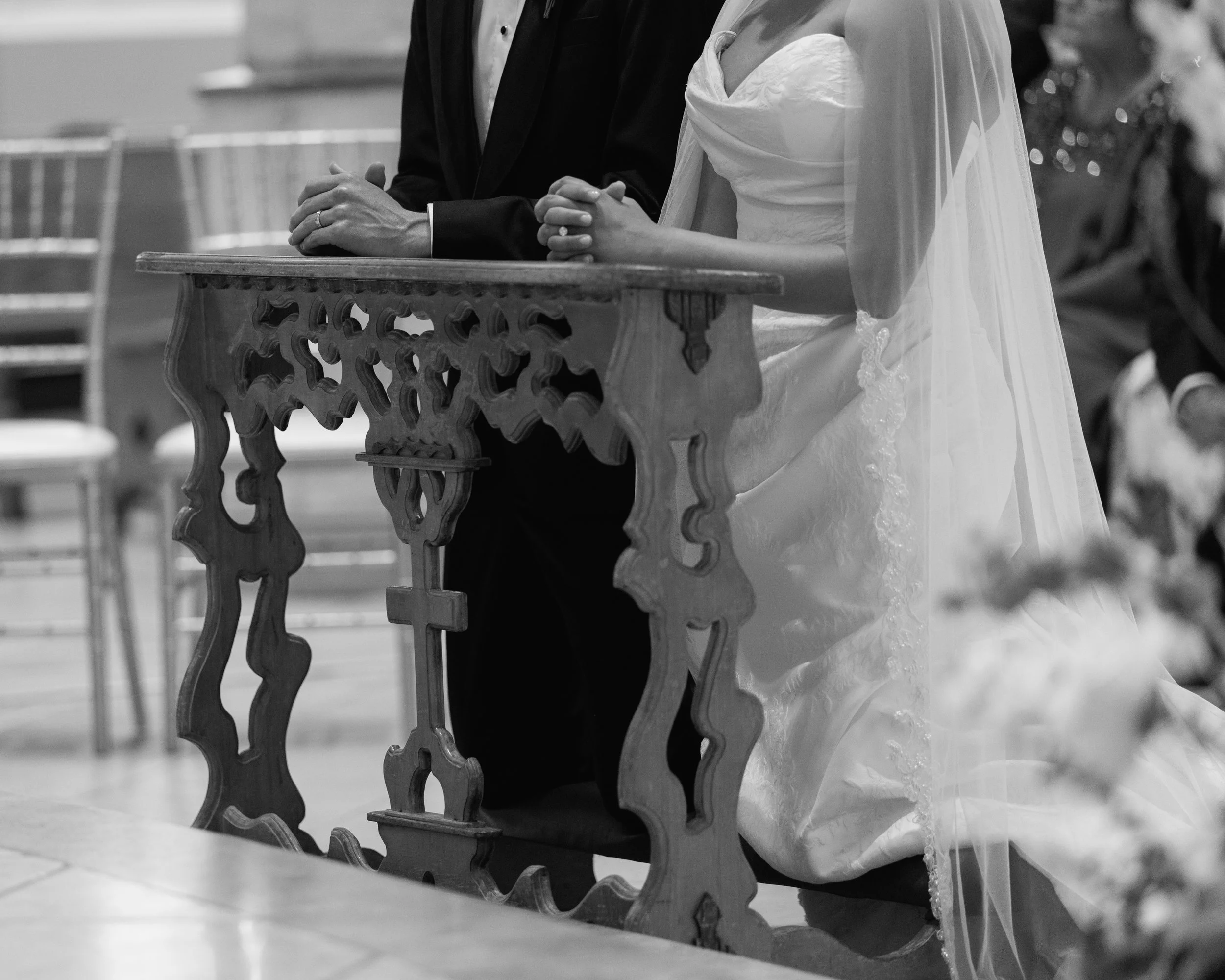 A black and white photo of a bride and groom standing at the altar, holding hands on a wooden pew during a wedding ceremony.
