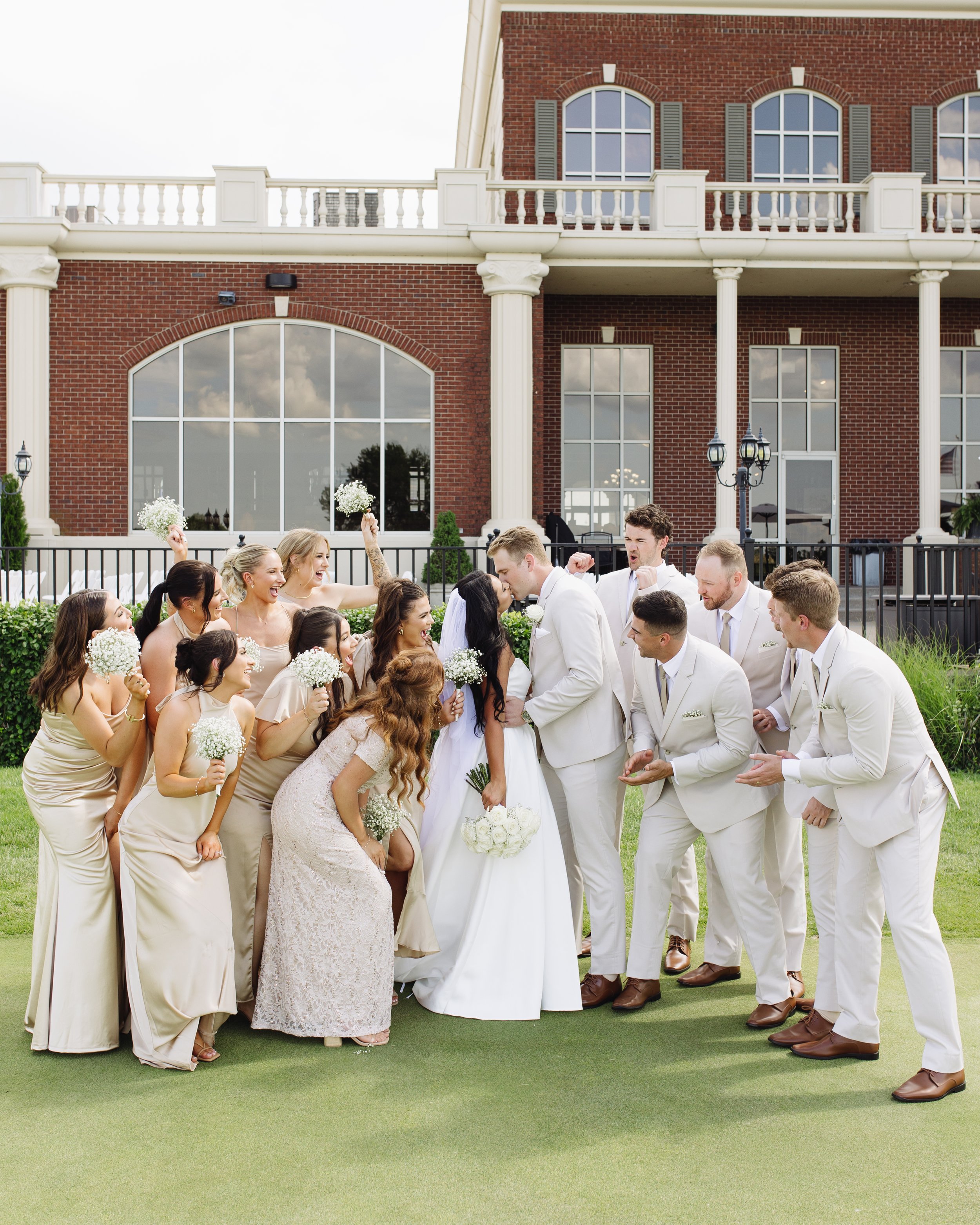 A group of wedding guests in formal attire standing outdoors on a green lawn, celebrating a wedding with a brick building with large windows in the background.