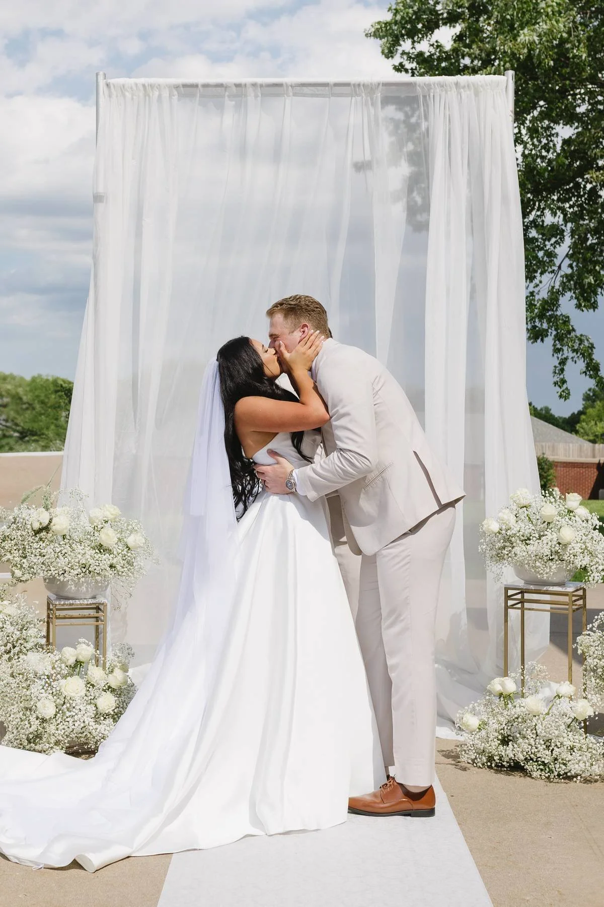 A newly married couple sharing a kiss during an outdoor wedding ceremony, with white drapery and floral arrangements of white roses and baby's breath behind them.