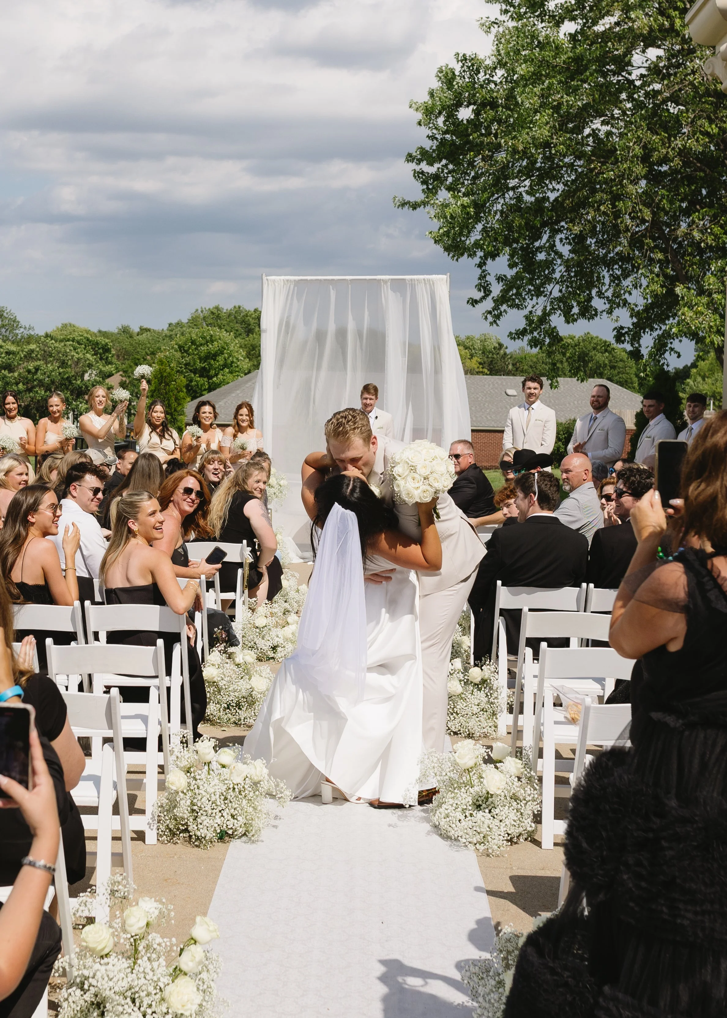 A wedding ceremony outdoors with guests seated on both sides of a white aisle. A couple is kissing at the altar, surrounded by floral arrangements of white flowers. Bridesmaids and groomsmen stand in the background, and there are trees and a building