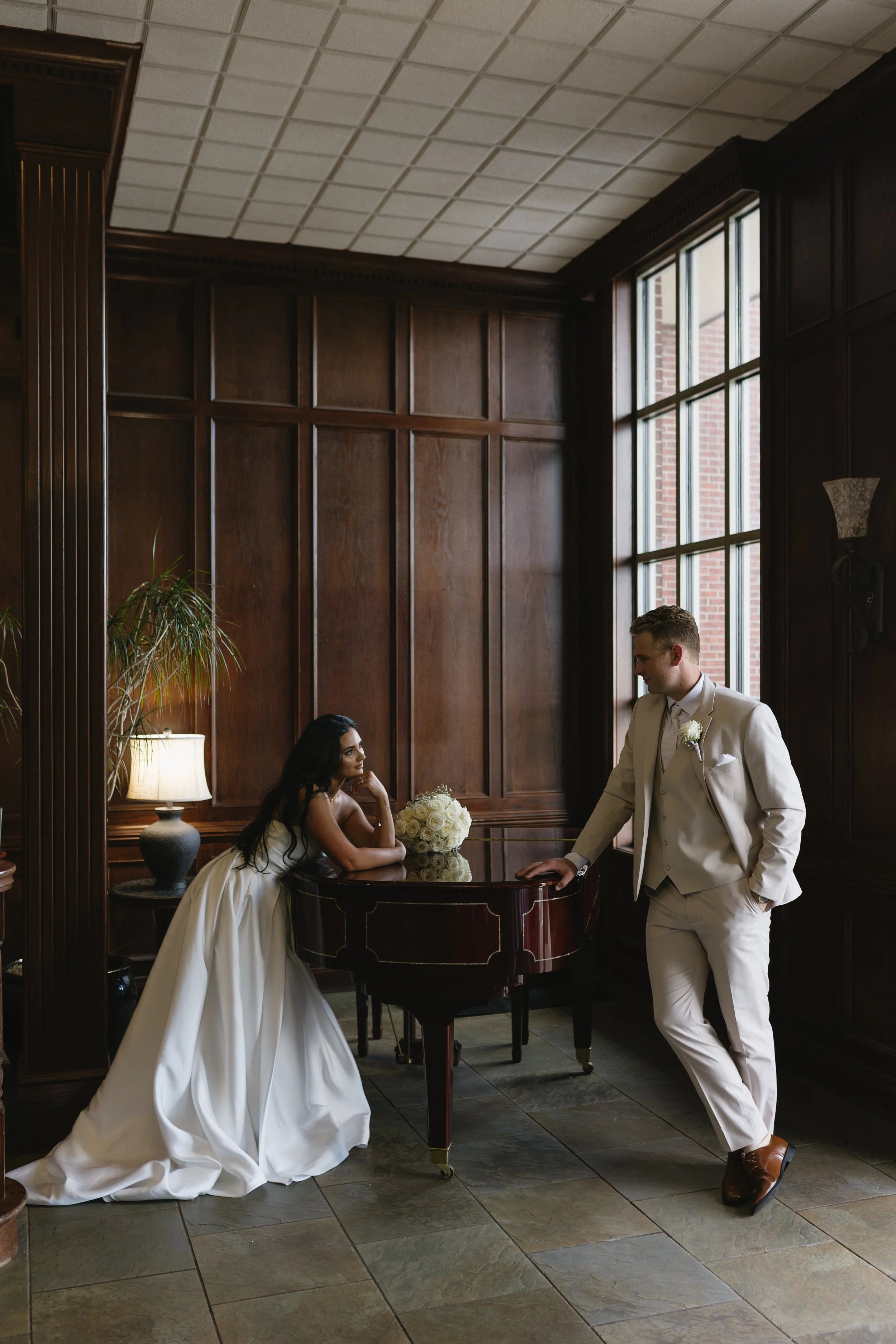 A woman in a white wedding gown sitting at a dark wood table with a bouquet of white roses, looking at a man in a white tuxedo. The man is standing with one hand resting on the table, looking at the woman. They are in a room with wood-paneled walls, 