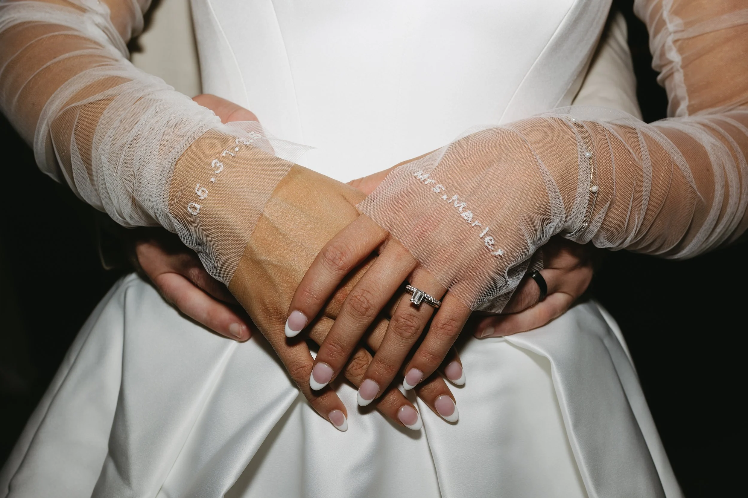 Close-up of a bride and groom holding hands, with the bride wearing a white wedding dress and sheer long sleeves with embroidery and pearls, and the groom in a white suit.