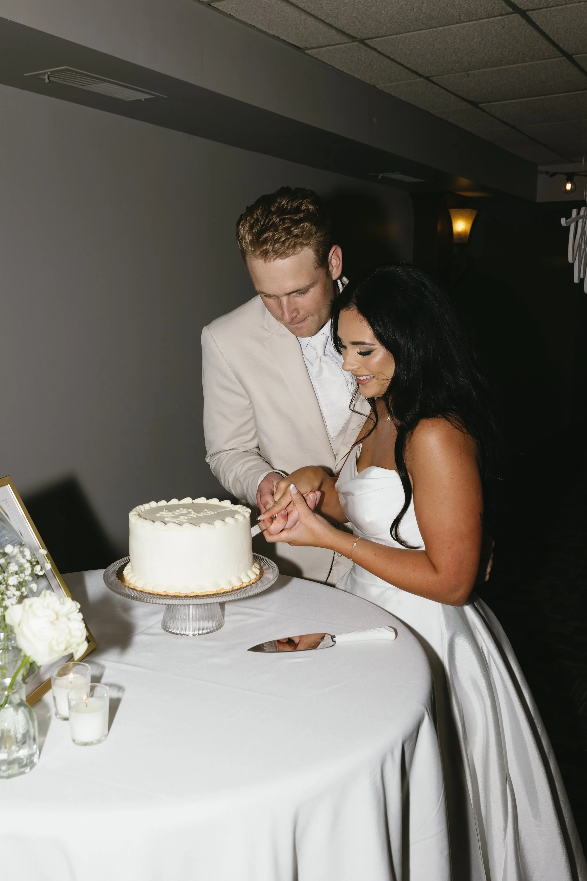 A couple, dressed in wedding attire, cutting a wedding cake at a reception table. The table is decorated with flowers and candles.