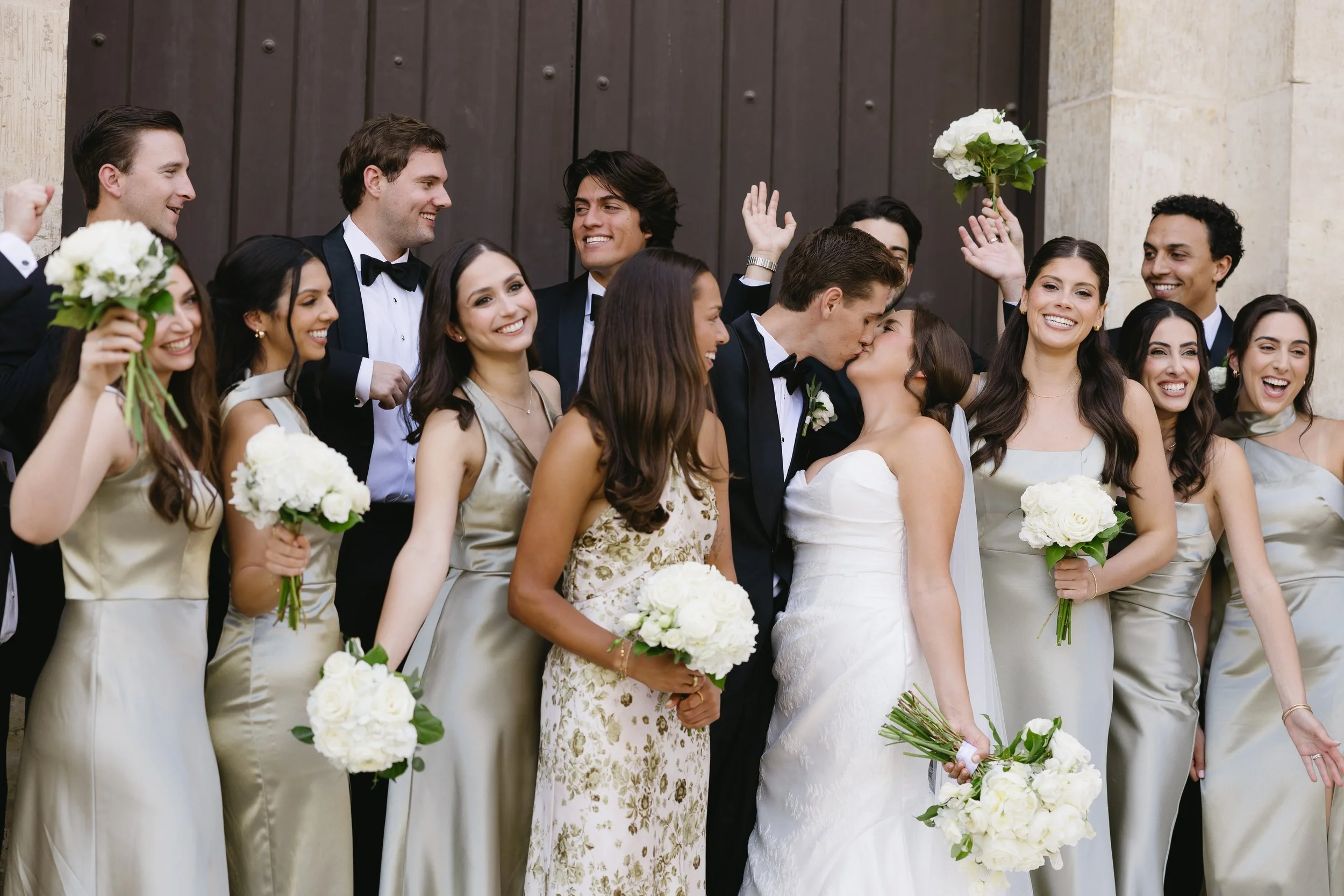 A group of wedding guests, including the bride and groom, celebrating outside in front of a dark wooden door. The bride is wearing a white wedding gown and holding a bouquet, while the groom is kissing her. The guests are dressed formally, with women