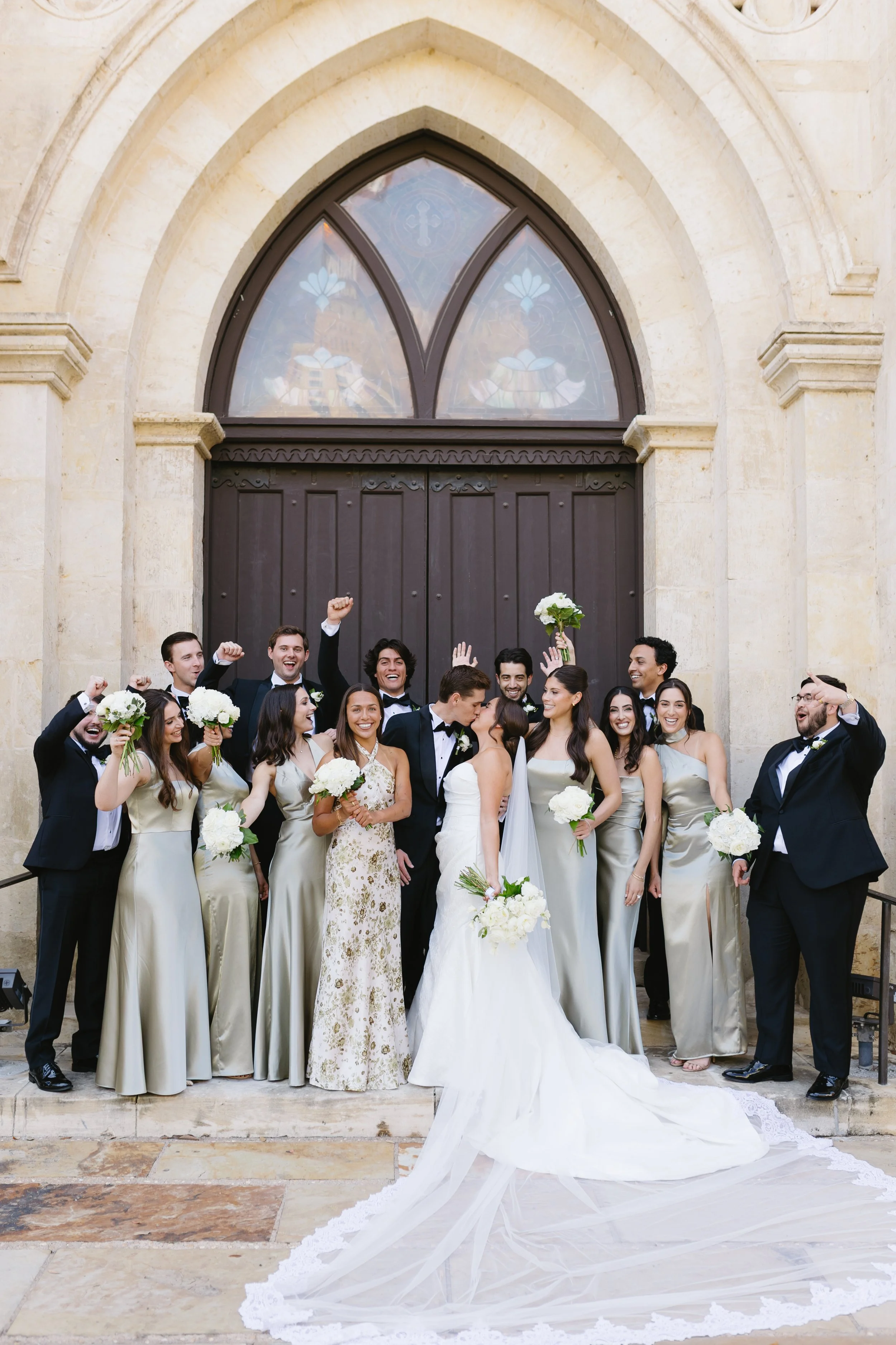 A group of people, dressed in formal attire, celebrating a wedding outside a church with a large wooden door and stained glass window. The bride and groom are kissing, surrounded by friends and family holding bouquets and raising their hands in celeb