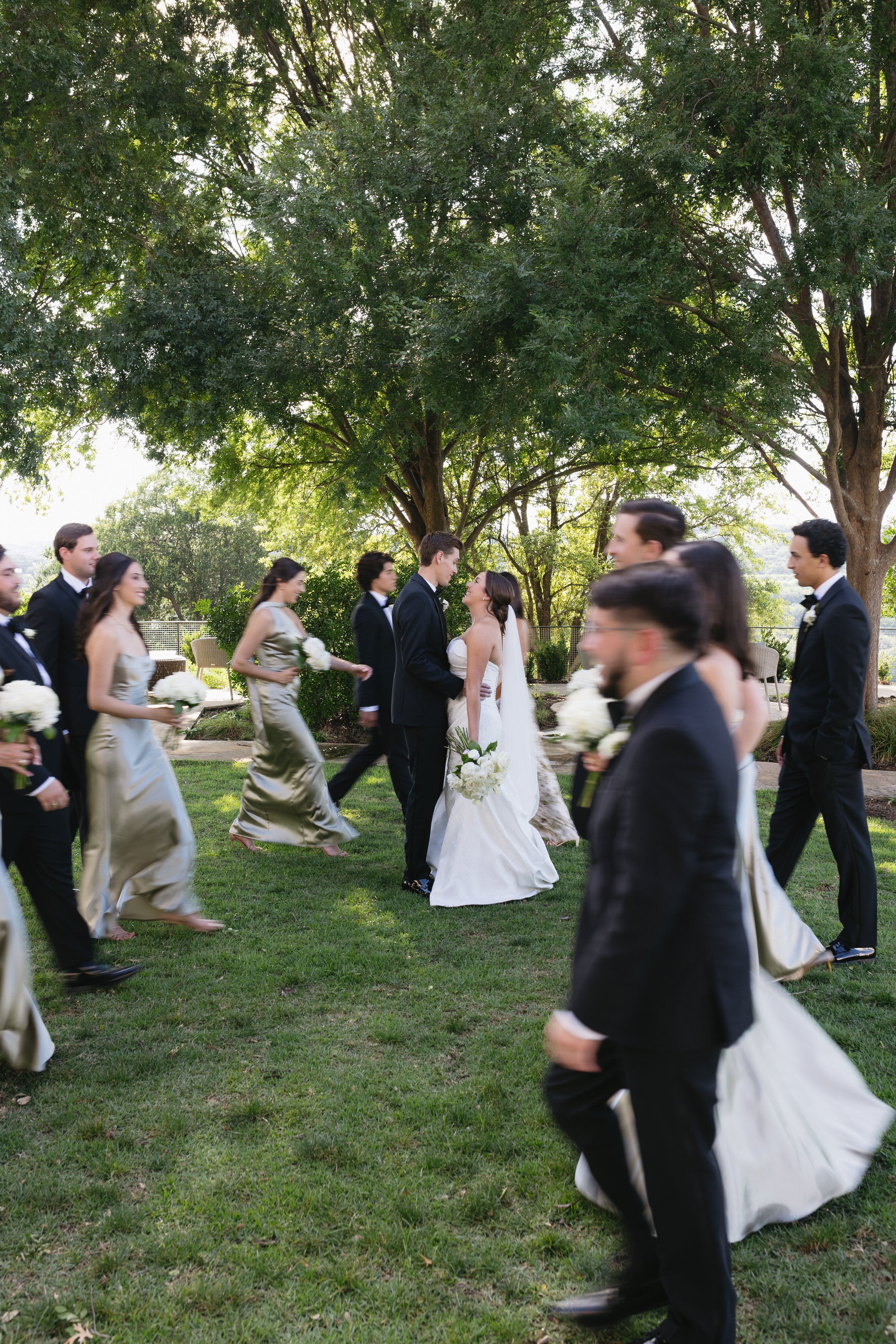 A wedding celebration taking place outdoors beneath a large green tree. The bride and groom are in the center, facing each other and holding hands. The guests, dressed in formal attire, are walking around the grassy area, some holding bouquets. The s