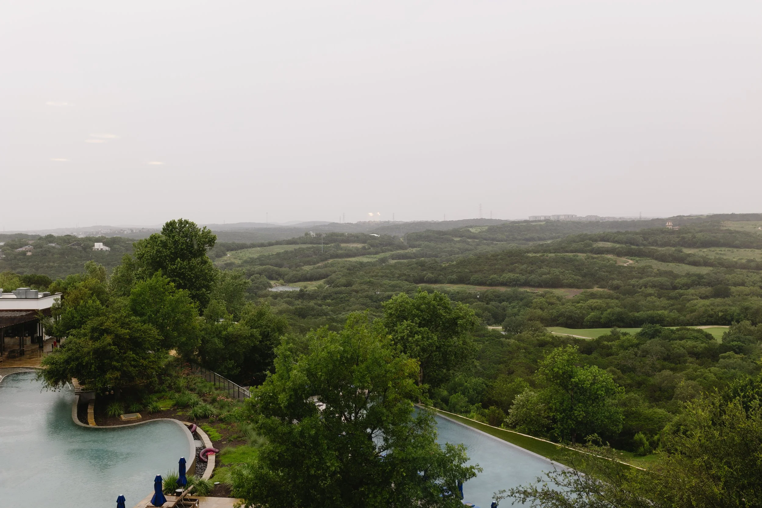 Scenic view of lush green trees, rolling hills, and distant buildings under a cloudy sky, with a swimming pool and umbrellas in the foreground.