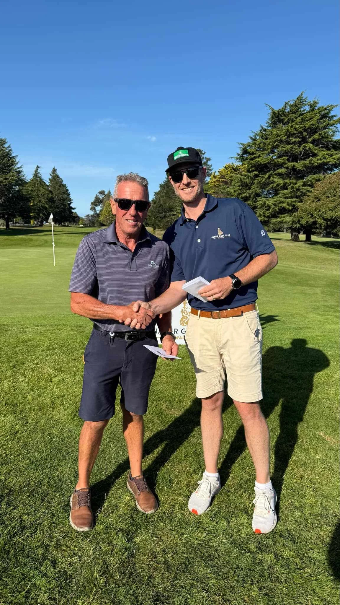 Two men shaking hands on a golf course, smiling and wearing sunglasses, with trees and a clear blue sky in the background.
