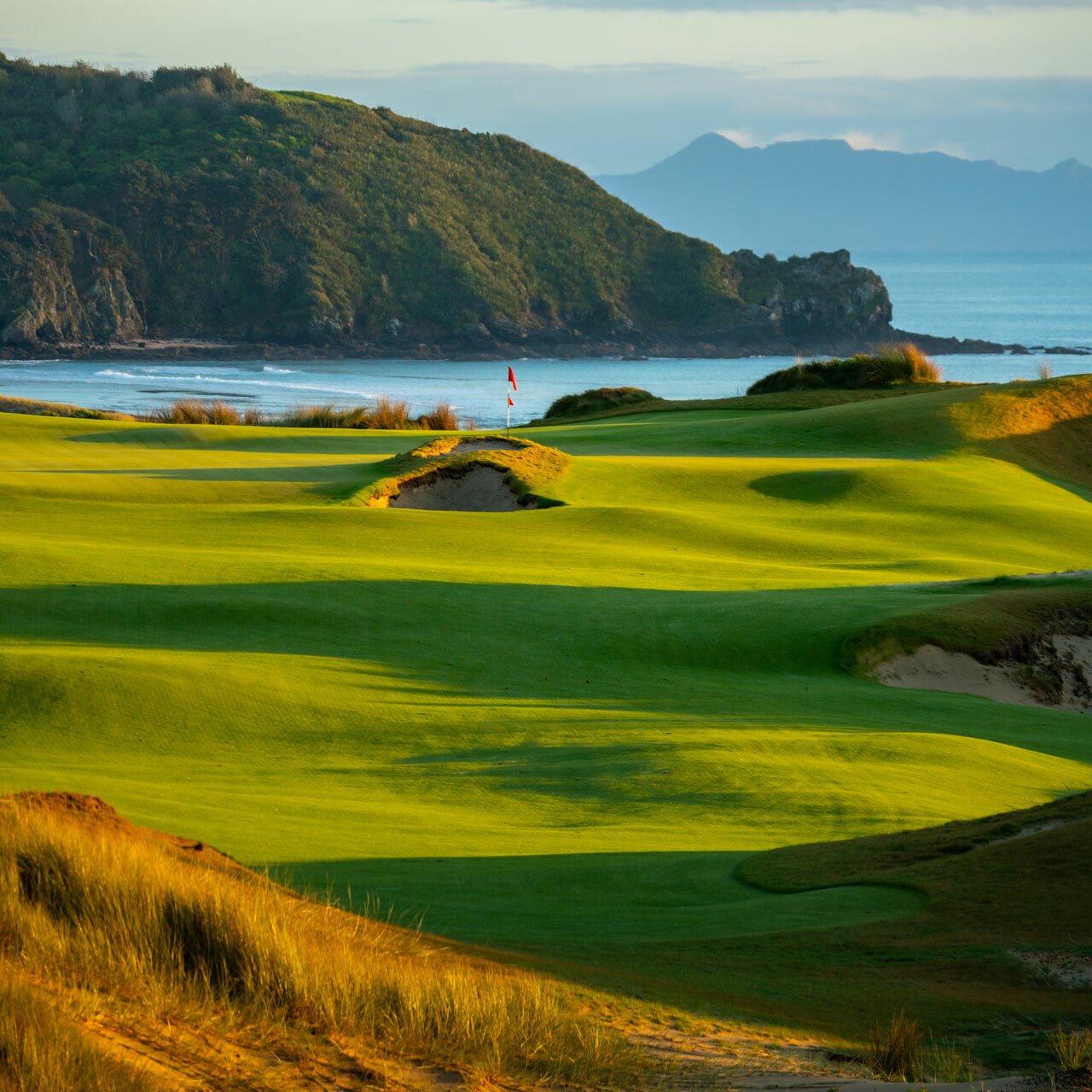 Scenic view of Te Arai golf course New Zealand with green fairways and a flag on the putting green near a coastline, with mountains and water in the background. Kiwi Golf Adventures
