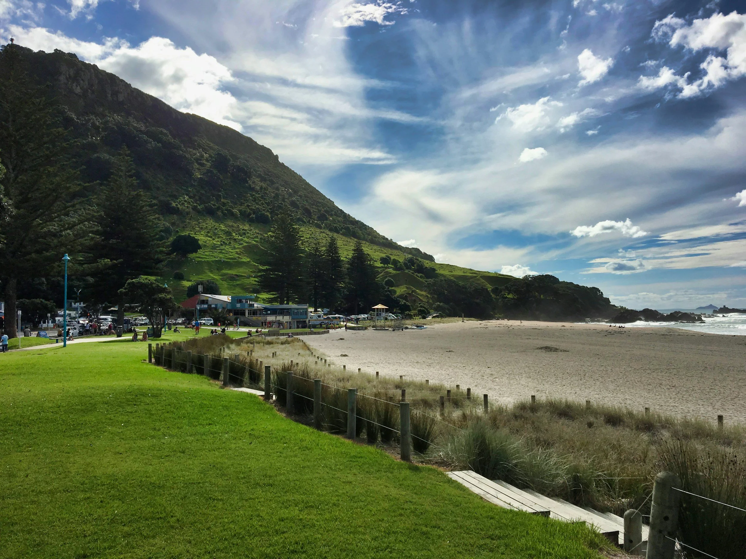 View of a grassy park near a beach with a mountain in the background under a partly cloudy sky. Mount Maunganui Beach - Kiwi Golf Adventures - Golf tours in new zealand