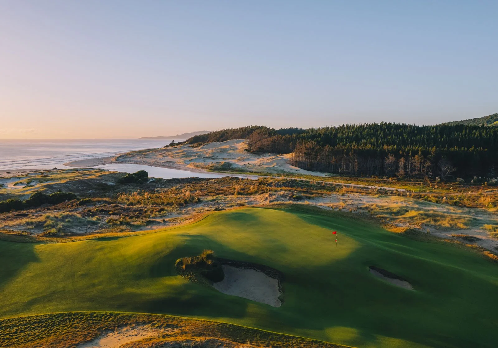 A scenic golf course near the coast of New Zealand - Te Arai South Course during sunset with green fairways, sand bunkers, and a red flag on the putting green, surrounded by dunes, trees, and ocean. Kiwi Golf Adventures