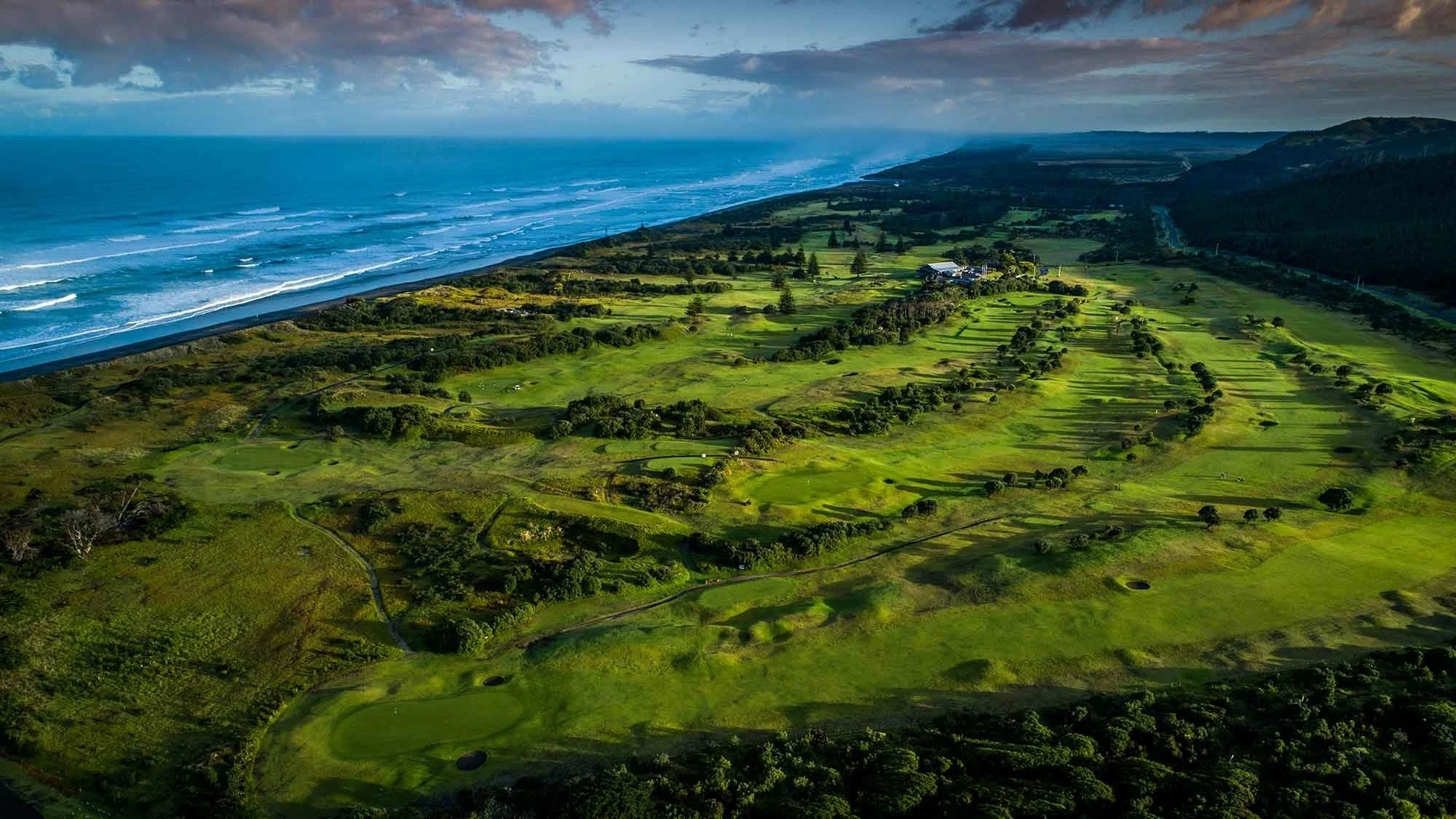 Aerial view of a lush green golf course along a coastline with waves and cloudy sky in the background. Muriwai Golf Course - Kiwi Golf Adventures - New Zealand Golf Tours