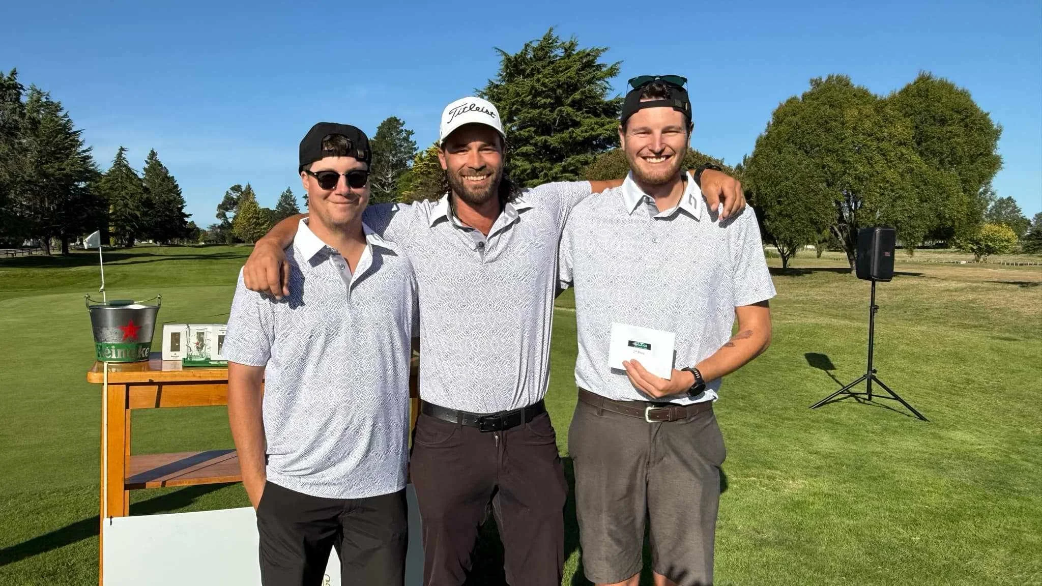 Three men standing arm-in-arm on a golf course, smiling, with a table and speaker in the background, during a sunny day. Kiwi Golf Adventures - Experience golf in New Zealand the Kiwi Way.