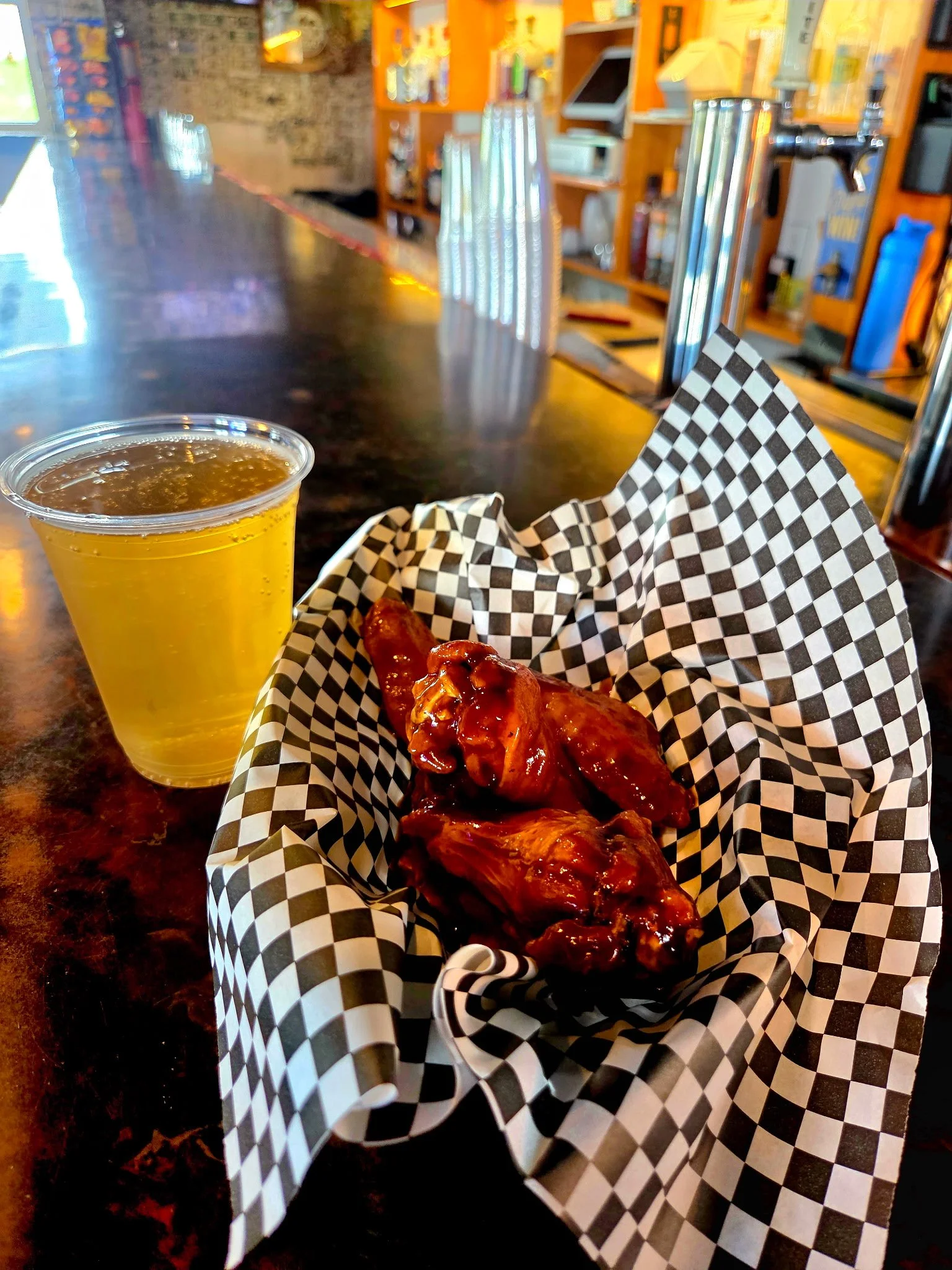 A plastic cup filled with beer, a basket with glazed chicken wings on checkered paper, and a counter with soda fountains and a rustic decor in the background.