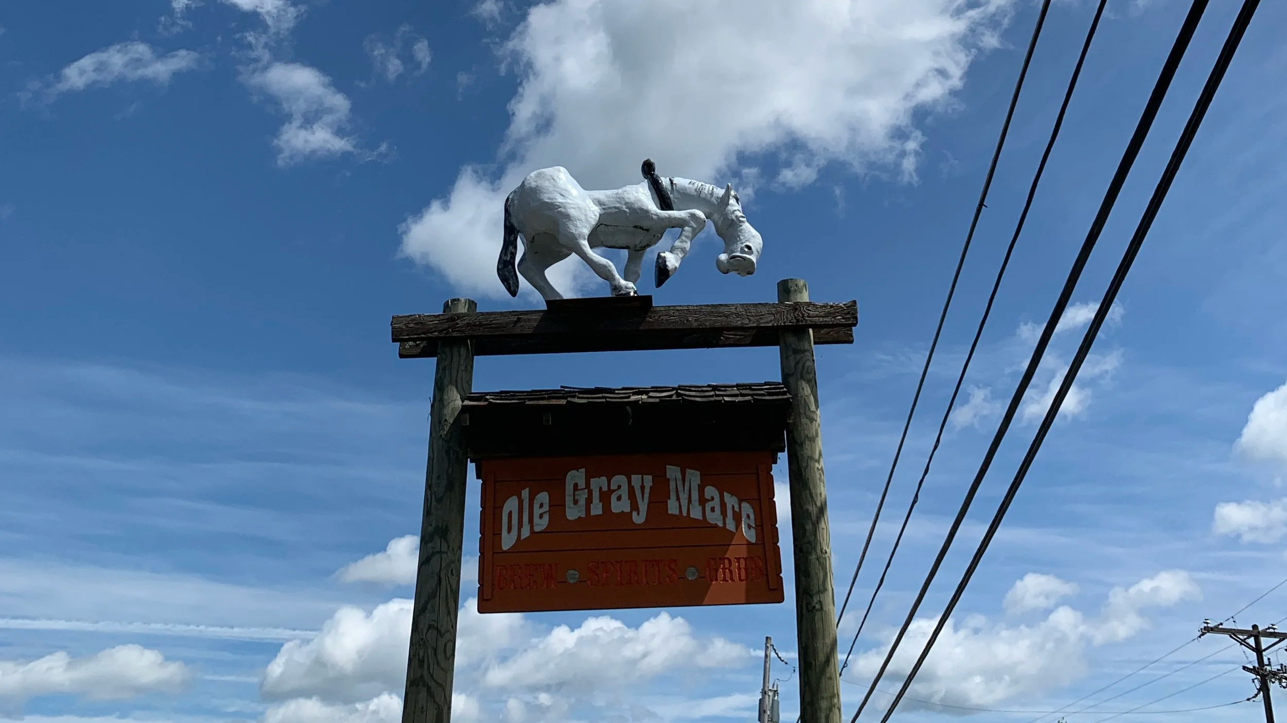 A weather vane in the shape of a white horse mounted on a wooden post against a partly cloudy sky, with power lines visible to the right.