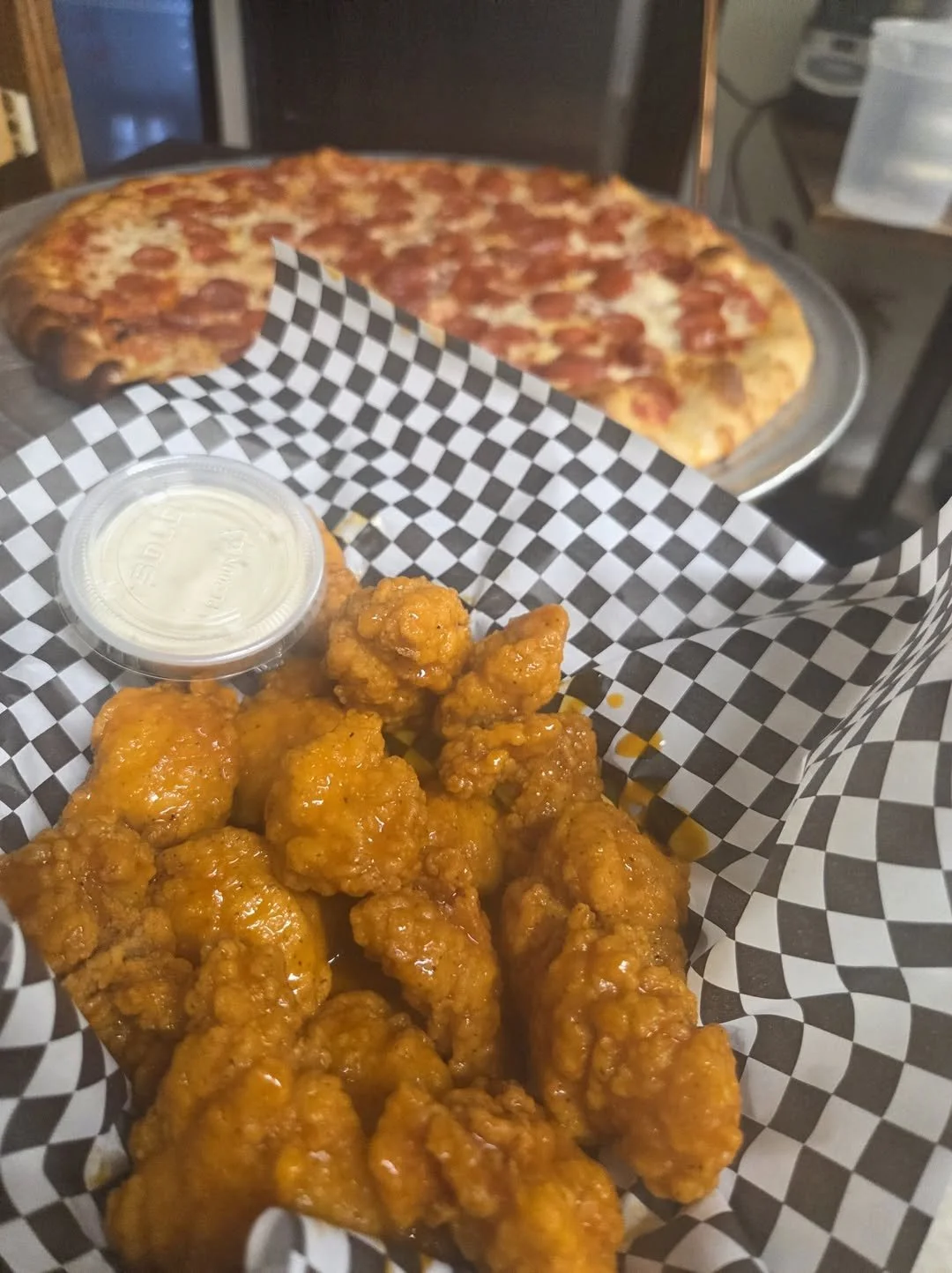 Basket of golden fried chicken pieces with a small container of dipping sauce, and a large pepperoni pizza in the background on a metal tray.