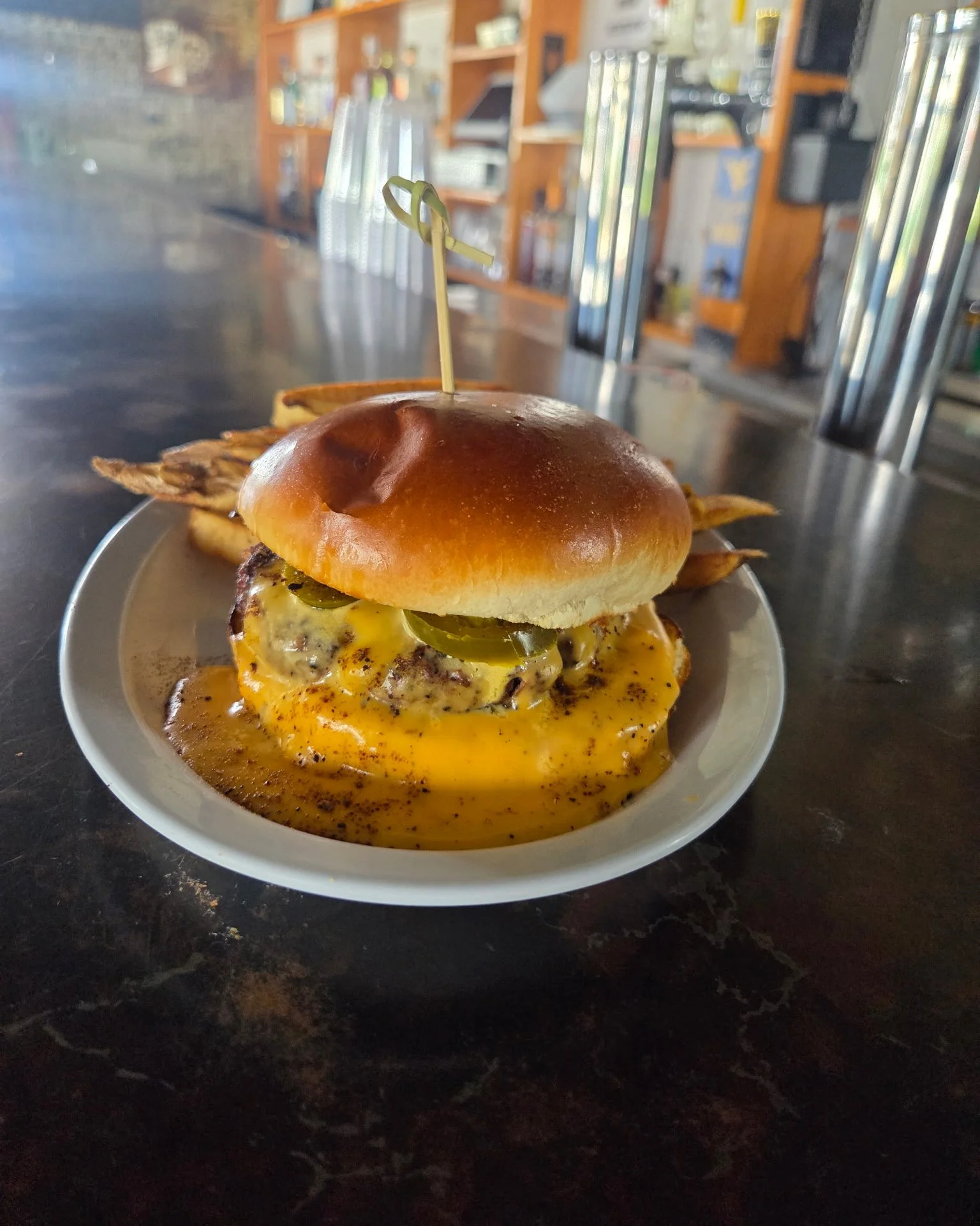 A burger with a shiny bun, yellow cheese, pickles, and crispy bacon on a plate, with spilled cheese sauce underneath, on a dark table in a restaurant setting.