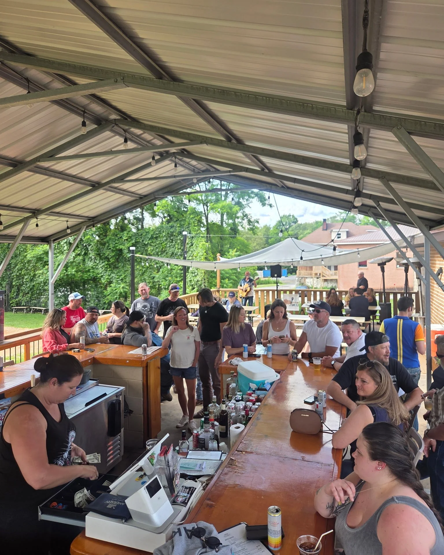 A large gathering of people under a metal pavilion, with some sitting at long wooden tables and others standing. There is a person at a cash register, with beverage cans and drinks on the tables. In the background, a person is playing guitar on the deck near green trees and a house.