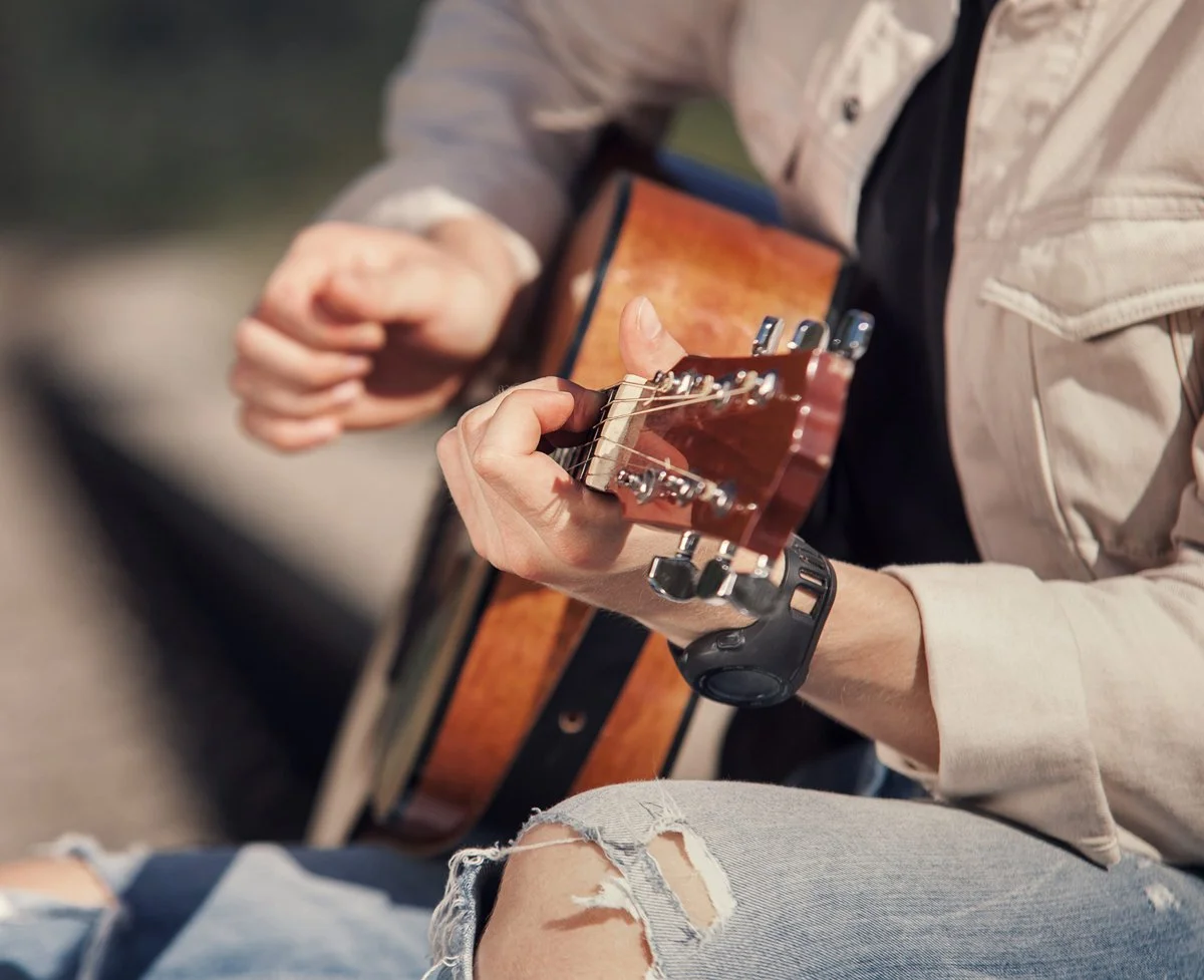 Person playing an acoustic guitar outdoors, sitting with ripped jeans.