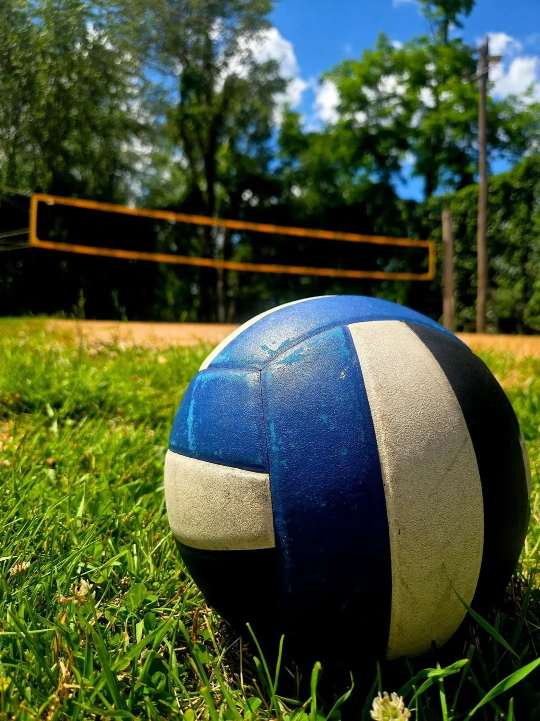 Close-up of a blue, white, and black volleyball on grass, with a volleyball court and net in the background under a blue sky with scattered clouds.