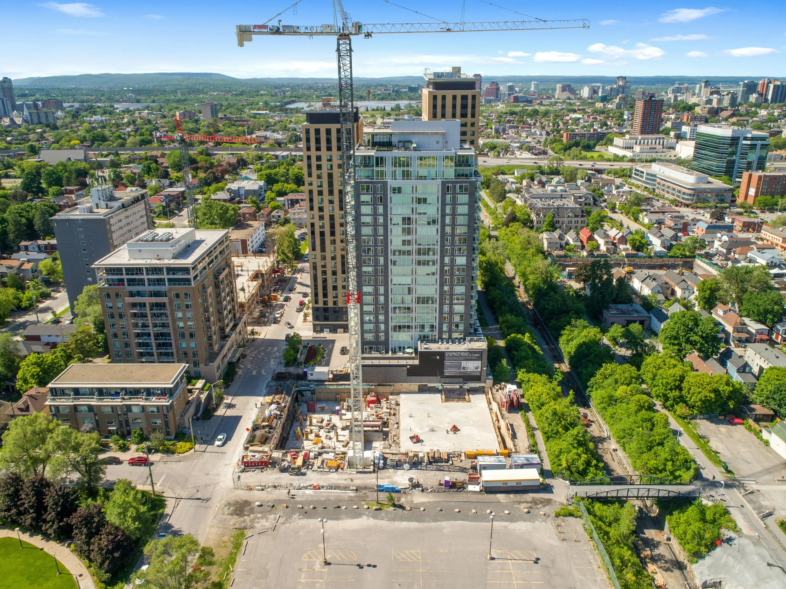 A tall building under construction with a large crane on top, located in an urban area with surrounding buildings, trees, and a distant city skyline.