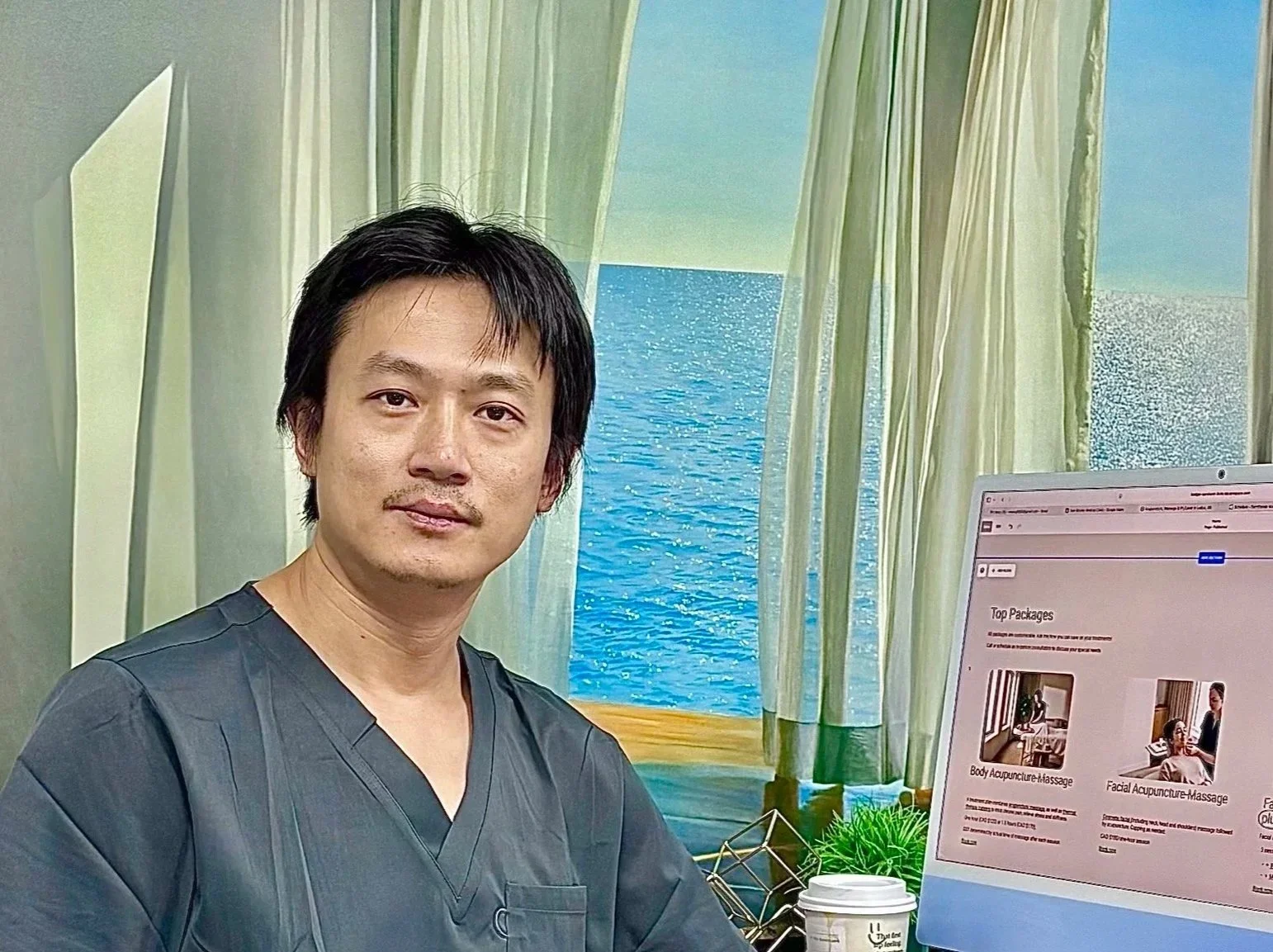A man with black hair and a mustache wearing dark scrubs sitting at a desk with a computer monitor displaying massage service options, with ocean-themed curtains and a window showing a blue ocean scene in the background.