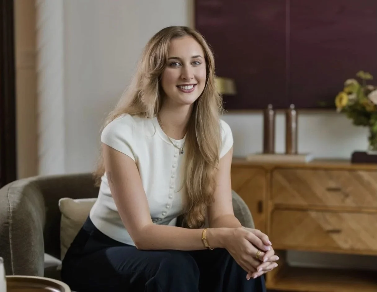 A woman with long, wavy blond hair and a white blouse sitting on a beige couch in a living room with a wooden sideboard and flower arrangement in the background, smiling at the camera.