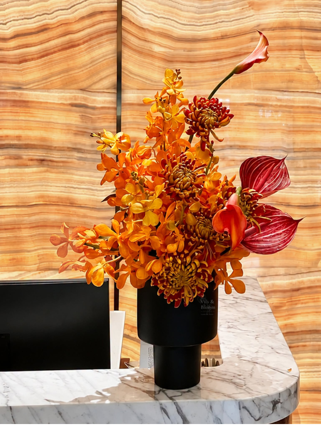 A floral arrangement with orange orchids, red and pink calla lilies, and red chrysanthemums in a black vase on a marble counter.