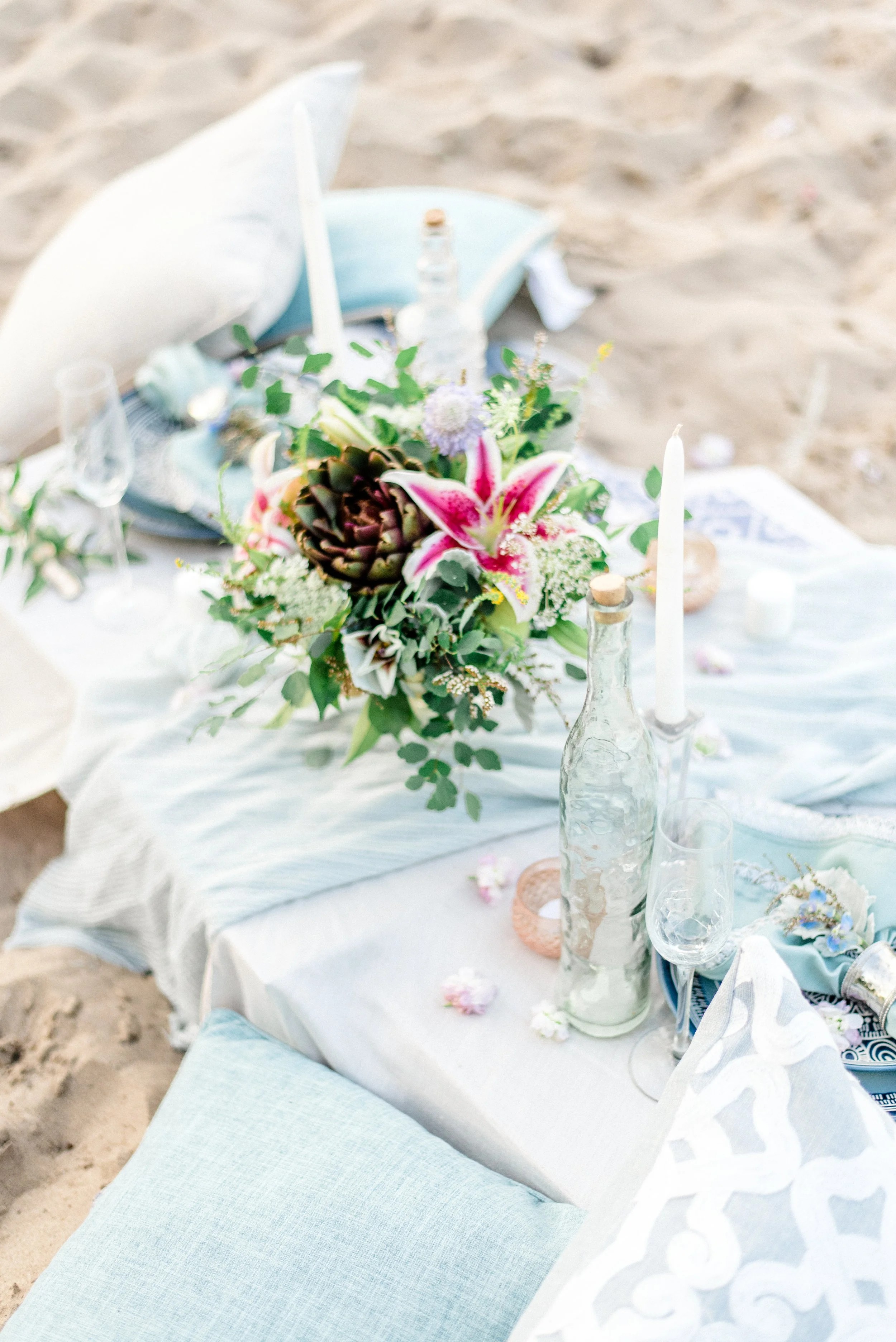 Beach wedding reception table with a floral centerpiece, candles, and glass bottles, set on white fabric on sandy ground.