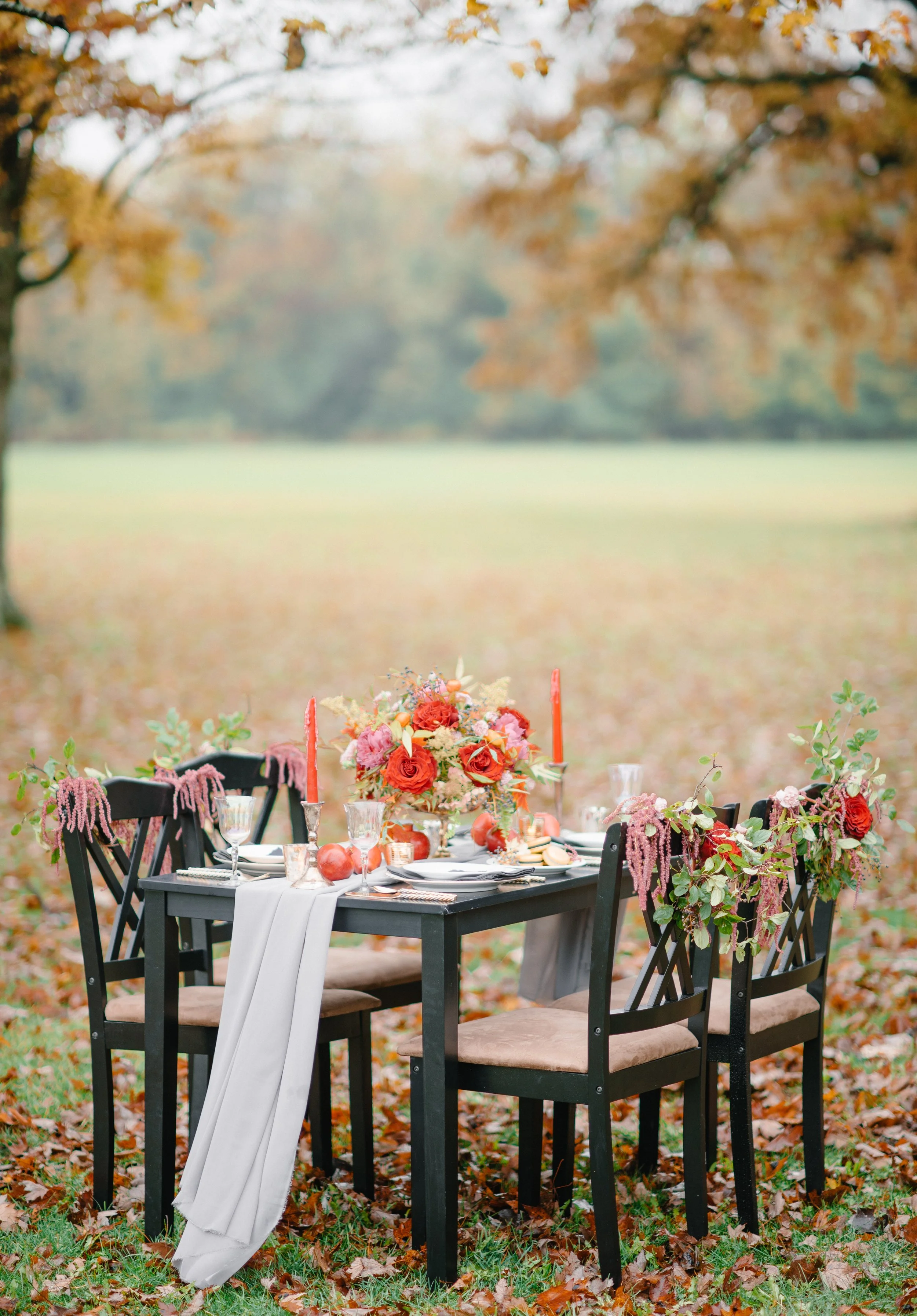 An outdoor dining table set with a floral centerpiece, candles, apples, and elegant tableware, surrounded by chairs, on a grassy field with fallen autumn leaves and trees with autumn foliage in the background.