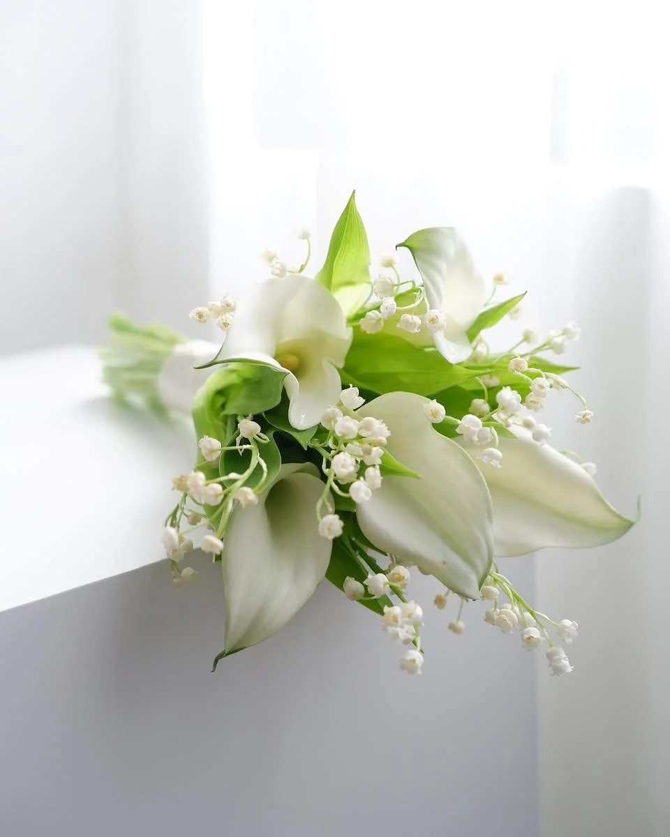 A bouquet of white calla lilies and lily of the valley flowers on a white surface with a bright background.