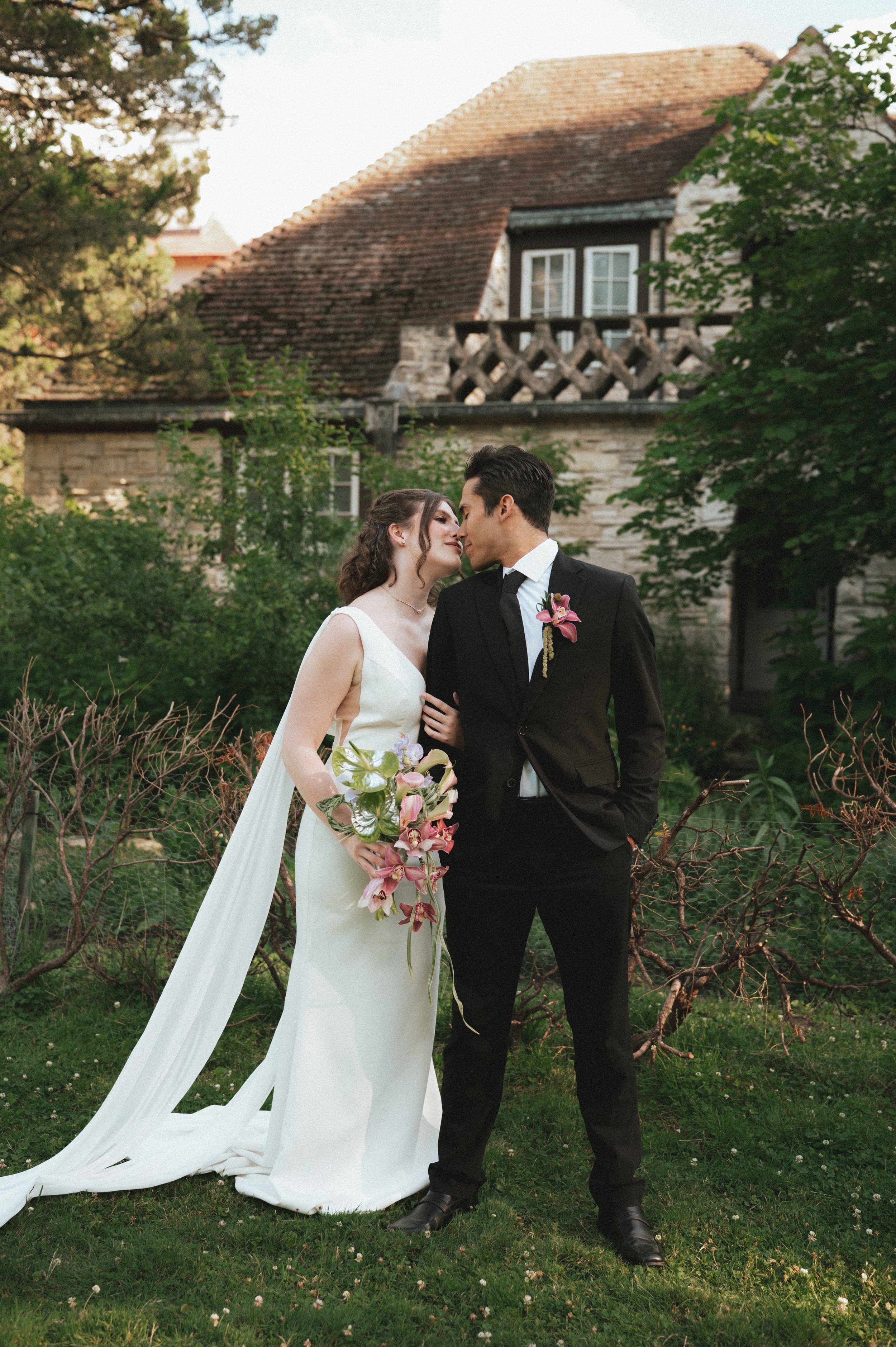 A bride and groom stand close to each other outdoors on a grassy lawn with trees and a stone house in the background. The bride wears a white wedding dress and holds a bouquet of pink and white flowers. The groom wears a black suit with a white shirt, black tie, and a pink boutonniere. They are leaning towards each other with their foreheads touching.