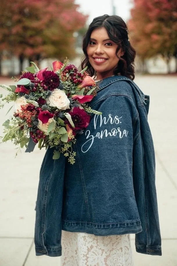 A woman with dark hair wearing a denim jacket with 'Mrs. Zamora' written on the back, holding a large bouquet of red, white, and pink flowers, standing outdoors with autumn trees in the background.