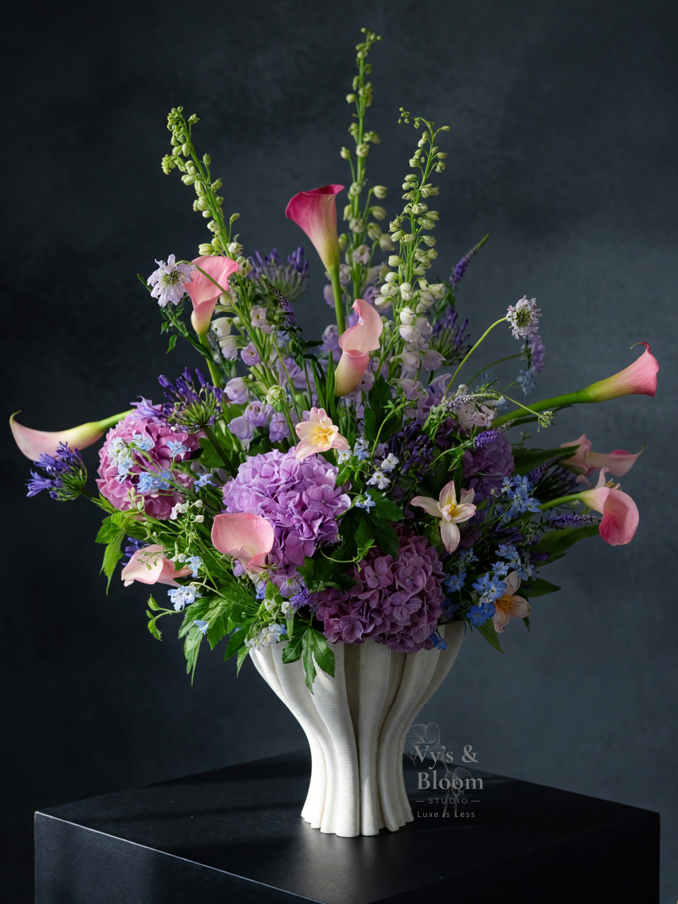 Elegant floral arrangement featuring pink calla lilies, purple hydrangeas, and other purple, white, and green flowers in a white ceramic vase with a striped pattern, placed on a black pedestal against a dark background.