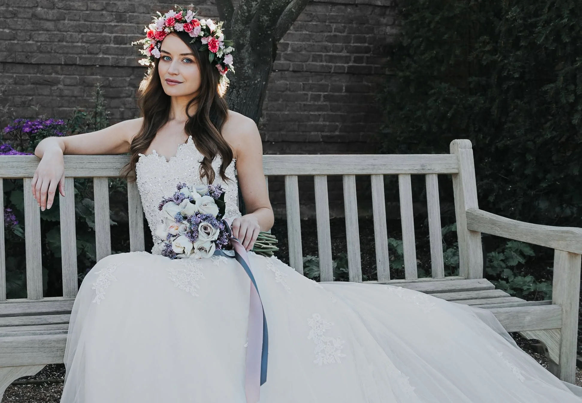 A young woman in a wedding dress sitting on a wooden park bench with a brick wall and green foliage in the background. She has long brown hair, light skin, blue eyes, and is wearing a floral crown with pink, white, and purple flowers, holding a bouquet of white calla lilies, purple flowers, and greenery.