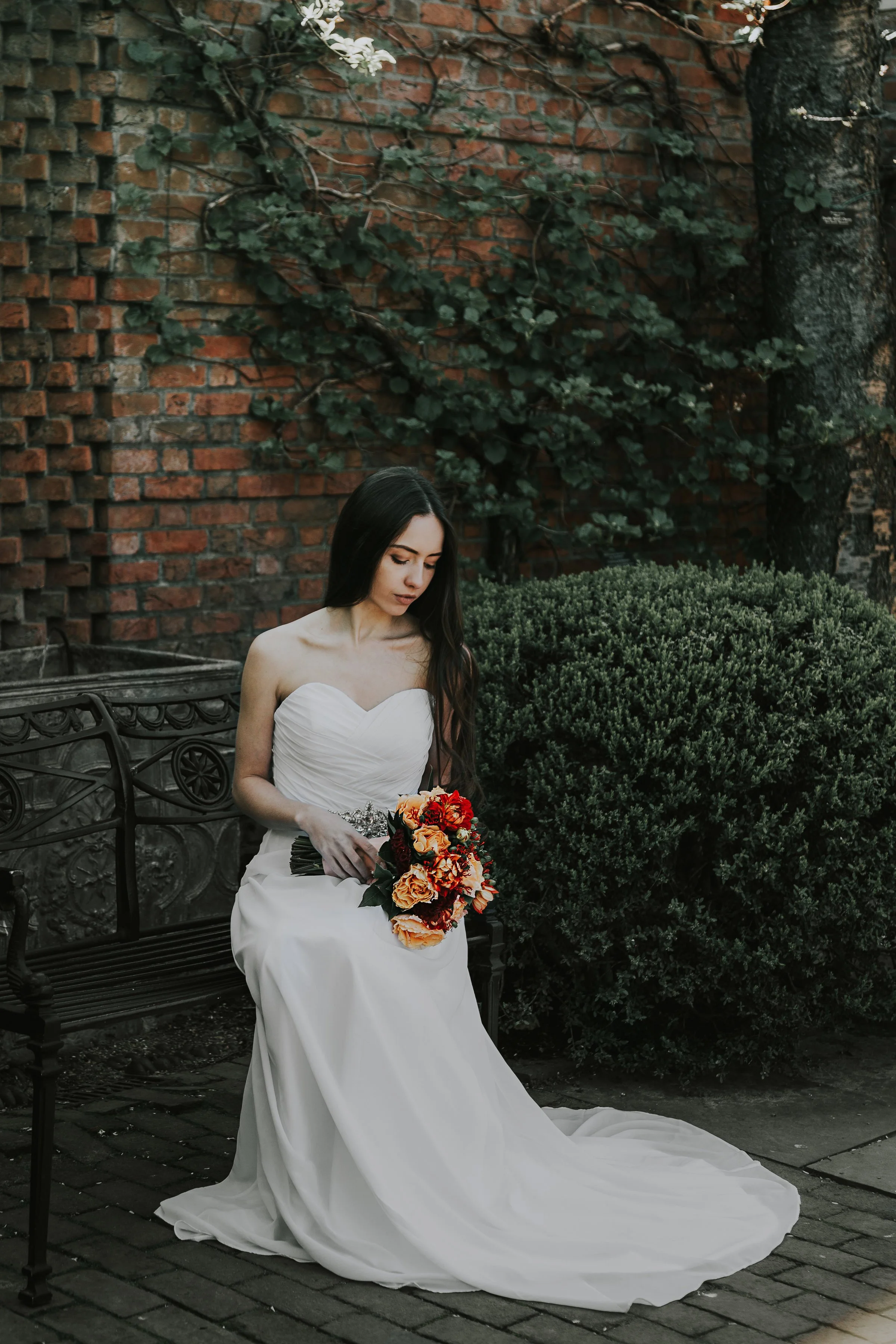 A woman in a white wedding dress sitting on a black metal park bench, holding a bouquet of orange and red flowers, with a brick wall and greenery in the background.