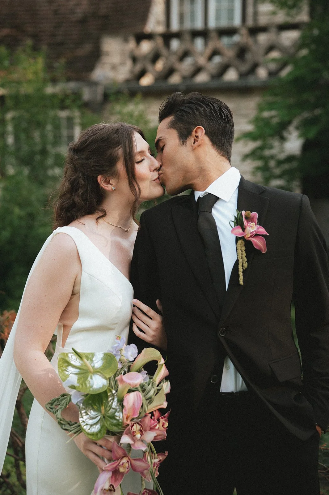 A bride and groom sharing a kiss outdoors, with greenery and a wooden structure in the background. The bride wears a white wedding dress and holds a bouquet of pink and purple flowers. The groom is dressed in a black suit with a boutonniere matching the bouquet.