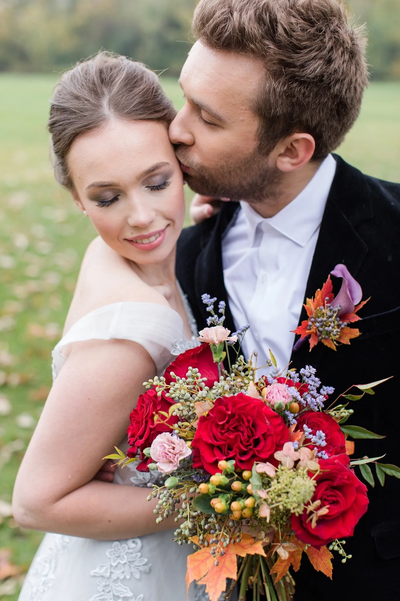 A bride and groom on their wedding day outdoors with the bride holding a large autumn flower bouquet with red, pink, purple, and orange flowers and foliage, both smiling.