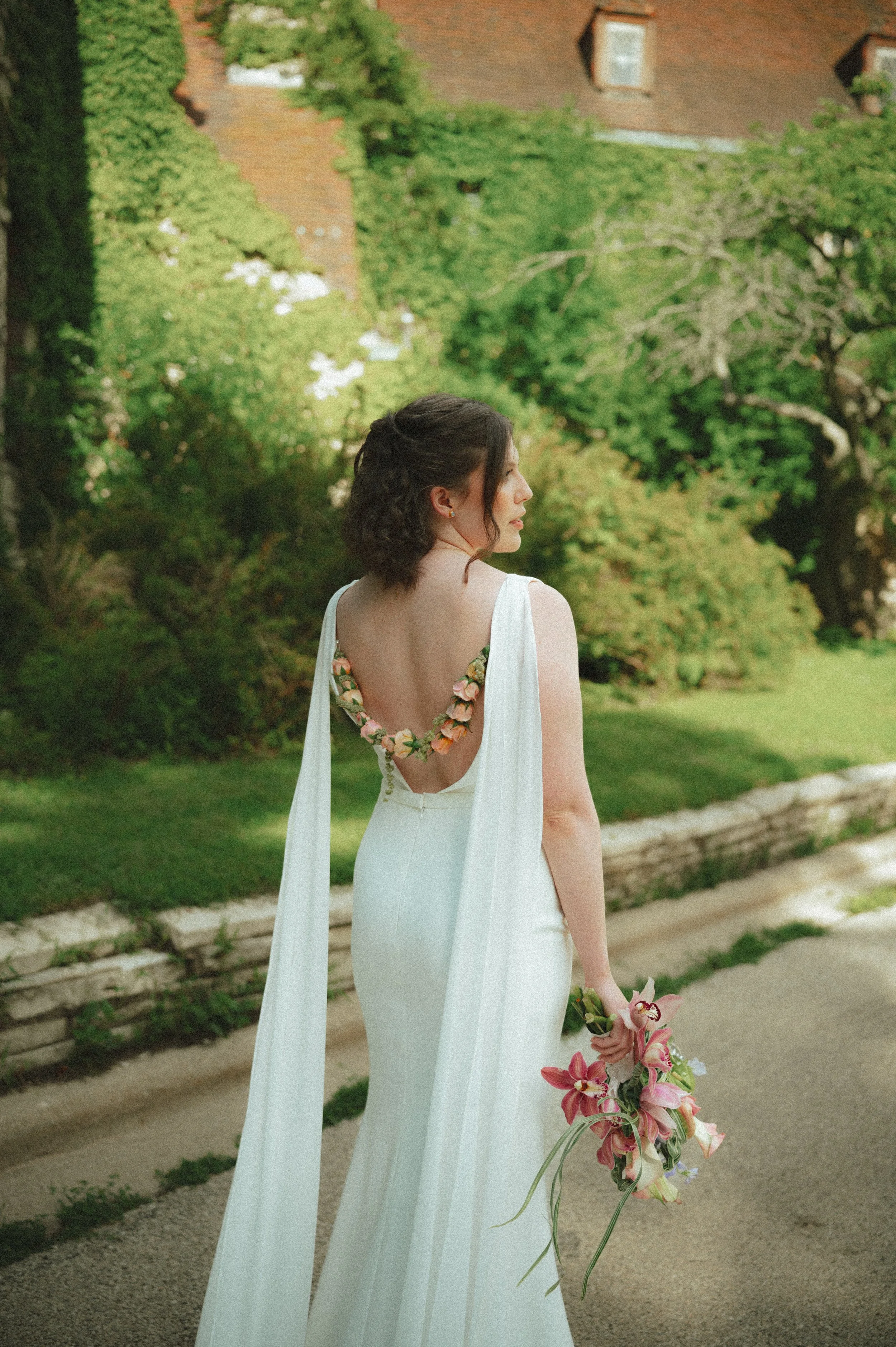 A woman in a white dress with floral details on the back, holding a bouquet of pink lilies, standing outdoors near a stone border with green trees and a brick house in the background.