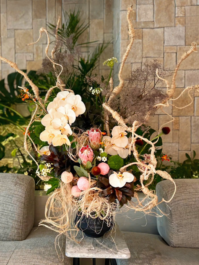 A decorative floral arrangement in a black pot featuring large white orchids, pink protea flowers, and curly, twisted branches, set on a small table between two gray couches, with a stone wall and green plants in the background.