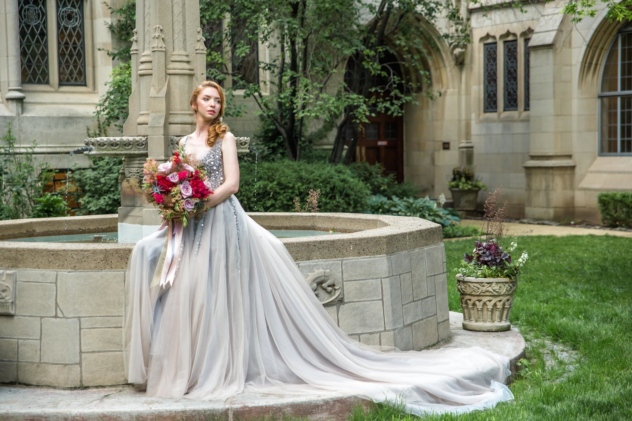 A woman in a silver and white gown holding a bouquet of pink and purple flowers, sitting on a stone fountain in a garden with historic stone buildings and trees in the background.