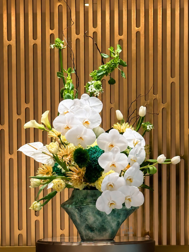 Elegant floral arrangement with white orchids, tulips, and other flowers in a green vase against a wooden wall background.
